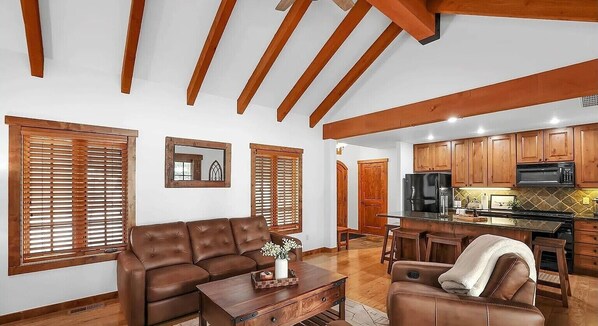 Un amplio salón con un sofá de cuero marrón, una mesa de centro de madera y un comedor con sillas de madera, además de una cocina al fondo.