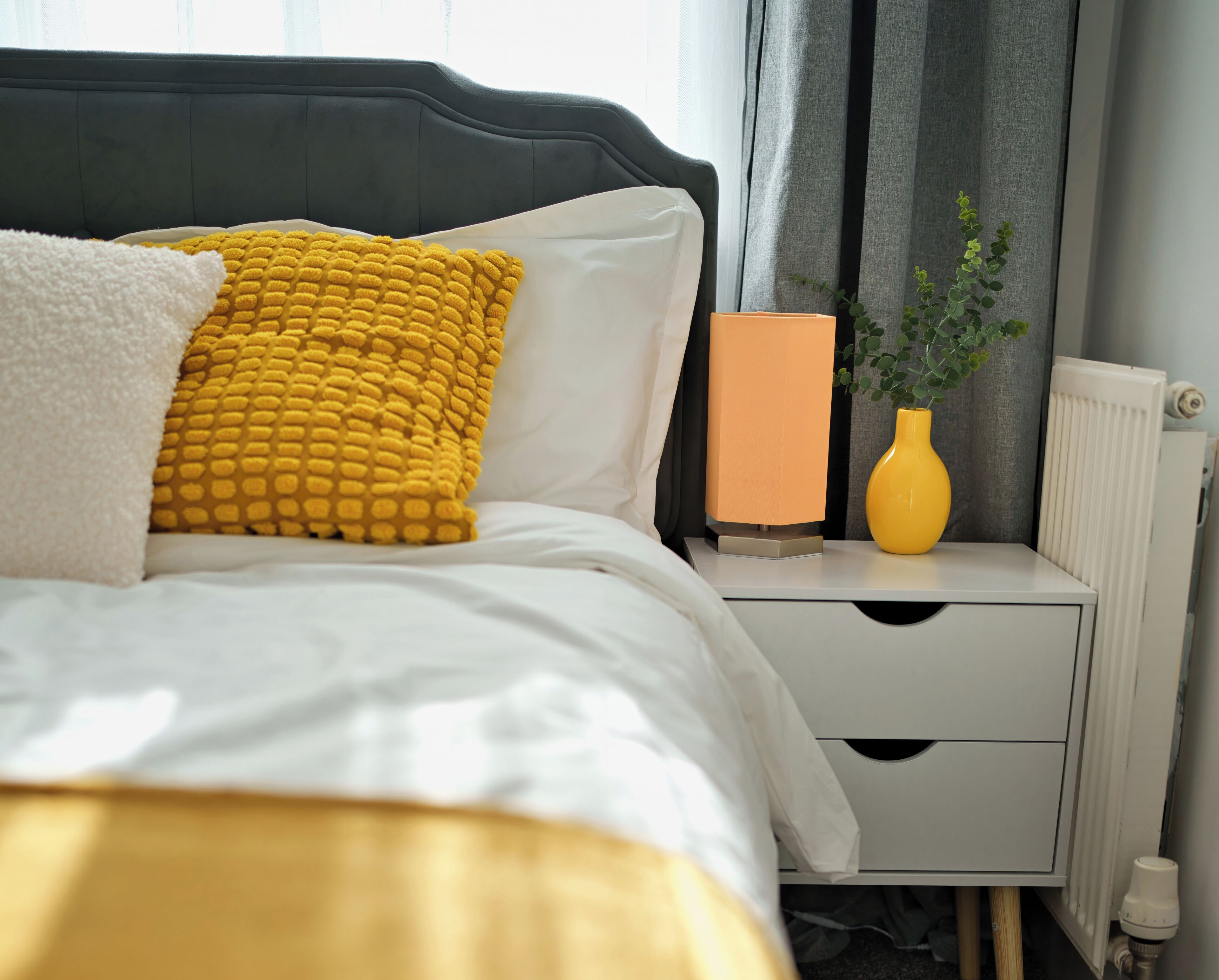 A neatly made bed with white and yellow pillows, a bedside table with a vase and a lamp, and a radiator.