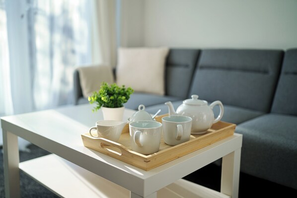 A tray with teacups and a teapot on a white table in a living room with a grey sofa and a plant in the background.