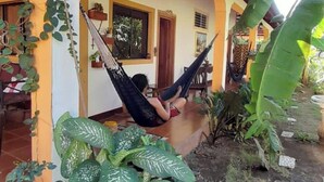 A person relaxing in a hammock on a porch with a view of a house and a garden. - RELAX IN WARM ROOMS AND SPACE TO REST COMFORTABLY. (León)