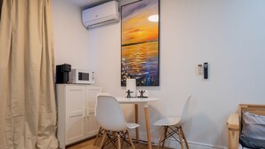 A small room with a white table, two white chairs, a wooden bed, a painting of a sunset, a microwave, and a remote control. - Spacious Studio for 4 | Full Kitchen + Laundry (Montréal)