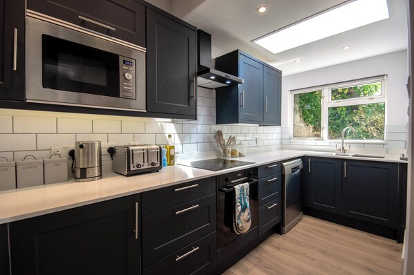 A modern kitchen with dark cabinets, stainless steel appliances, and a window providing natural light. - Stylish Salcombe House Walk to Beach & Town, Pets, Garden, 2 Parking, Sleeps 6 (Salcombe)