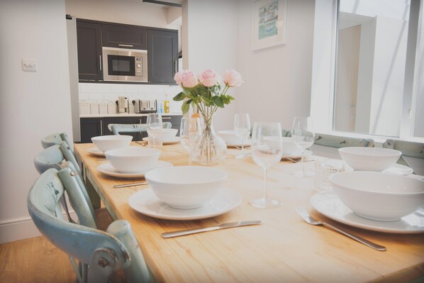 A dining area with a wooden table set for six, white bowls, wine glasses, and a vase with flowers. - Stylish Salcombe House Walk to Beach & Town, Pets, Garden, 2 Parking, Sleeps 6 (Salcombe)