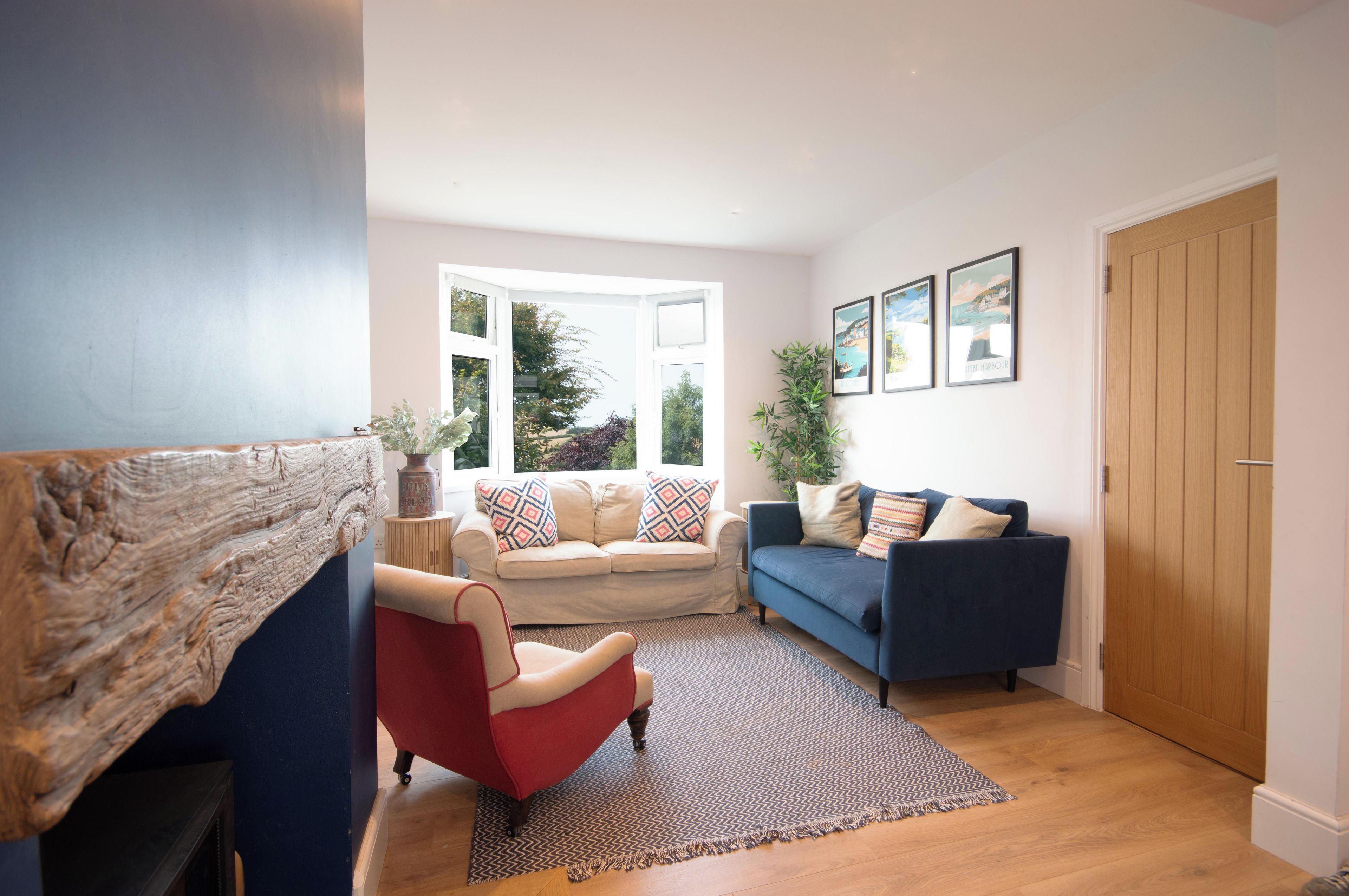 Een woonkamer met een rode fauteuil, een blauwe bank en een beige bank met gekleurde kussens. Er is een groot raam met uitzicht op groen.