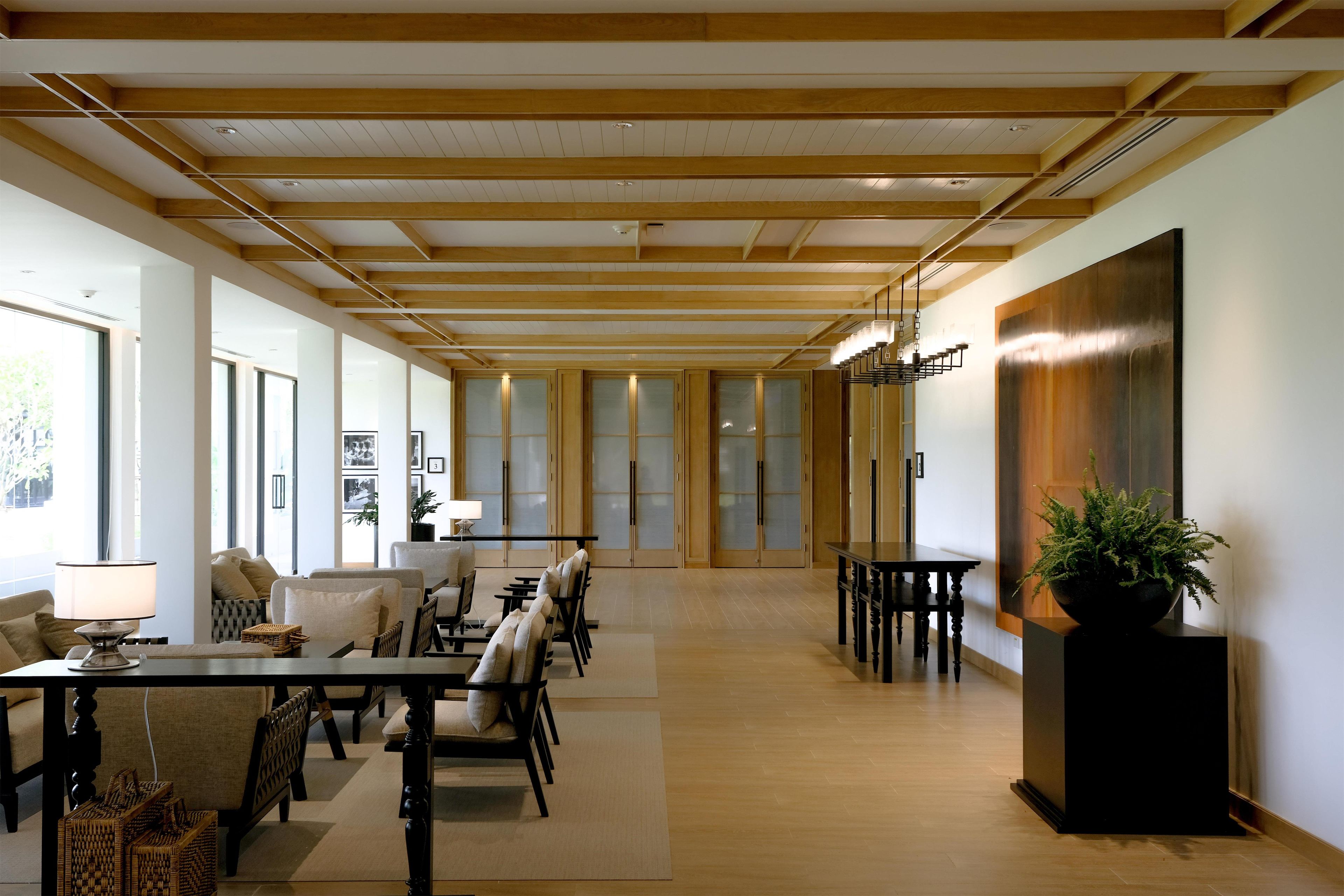 A spacious room with a high ceiling, wooden beams, a dining area with chairs and tables, and large windows allowing natural light.