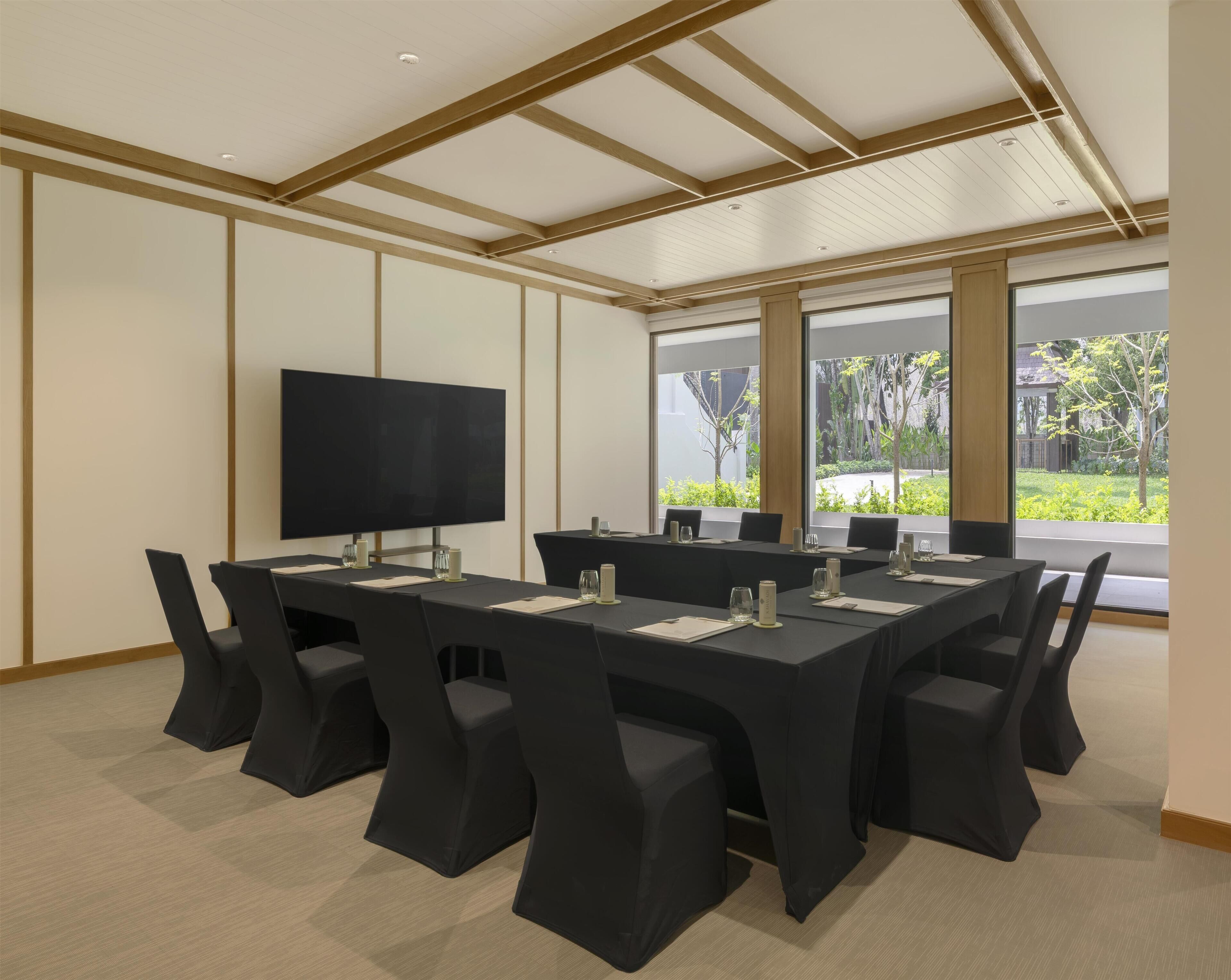 Une salle de conférence équipée d’un grand téléviseur à écran plat, de plusieurs tables avec des chaises et de grandes fenêtres donnant sur un paysage verdoyant.