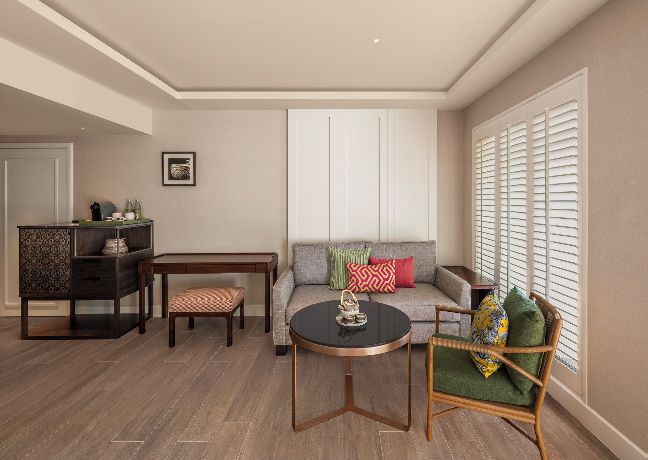 Un salon comprenant un canapé, une table ronde, une chaise en bois, un petit meuble et une fenêtre avec des stores.