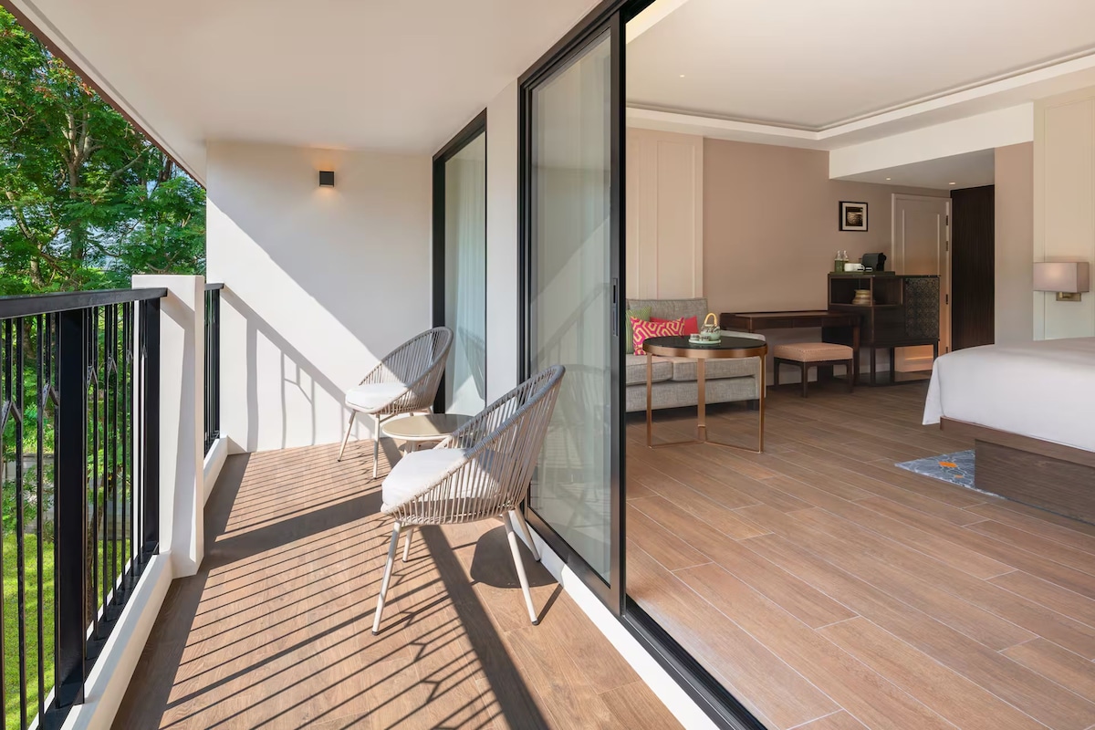 A modern hotel room with a balcony, a dining area, and a view of greenery.