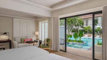 A hotel room with a large bed, a sofa, a chair, a small table, a lamp, and a view of an outdoor swimming pool.
