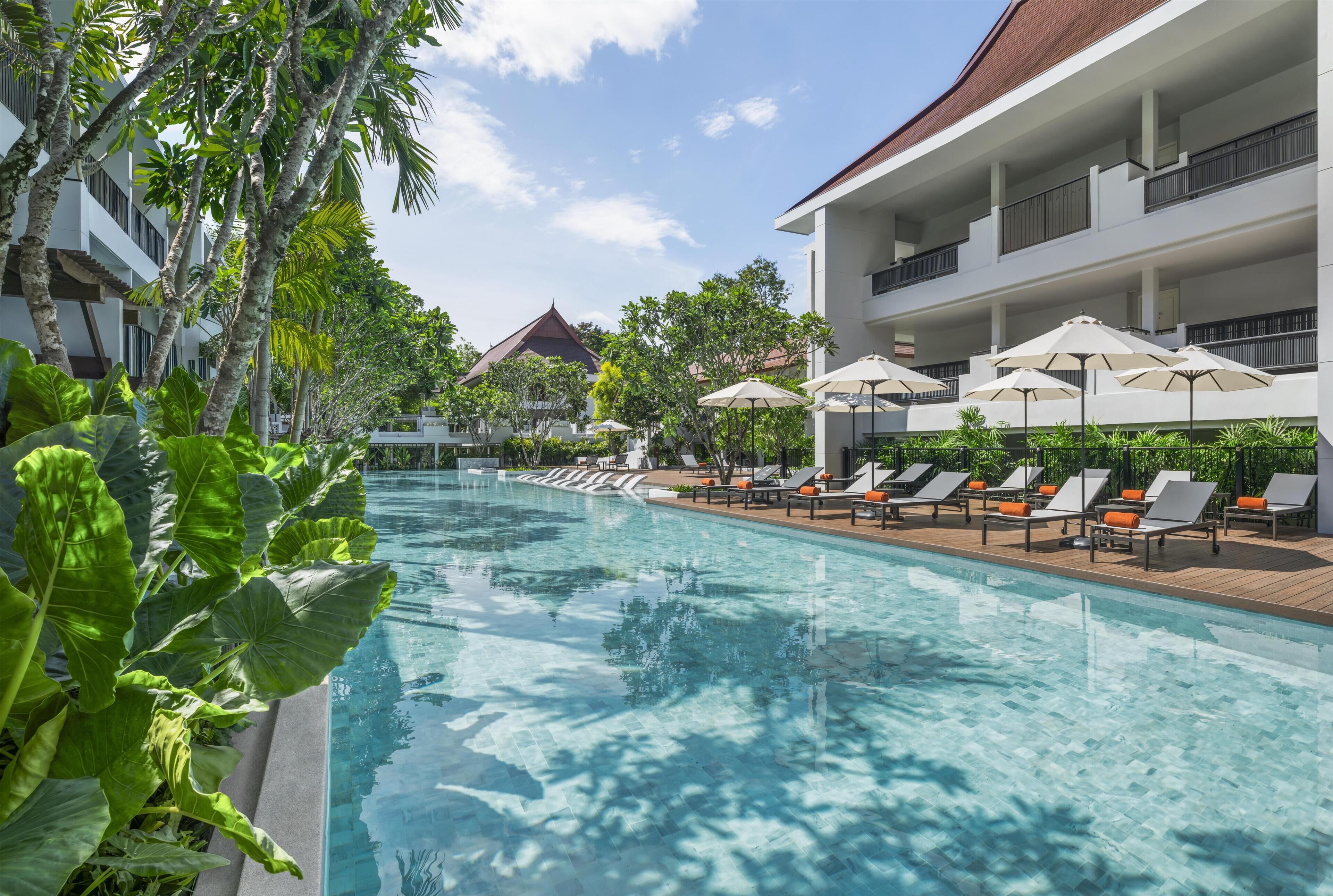 A swimming pool with lounge chairs and umbrellas, surrounded by greenery and a multi-story building.