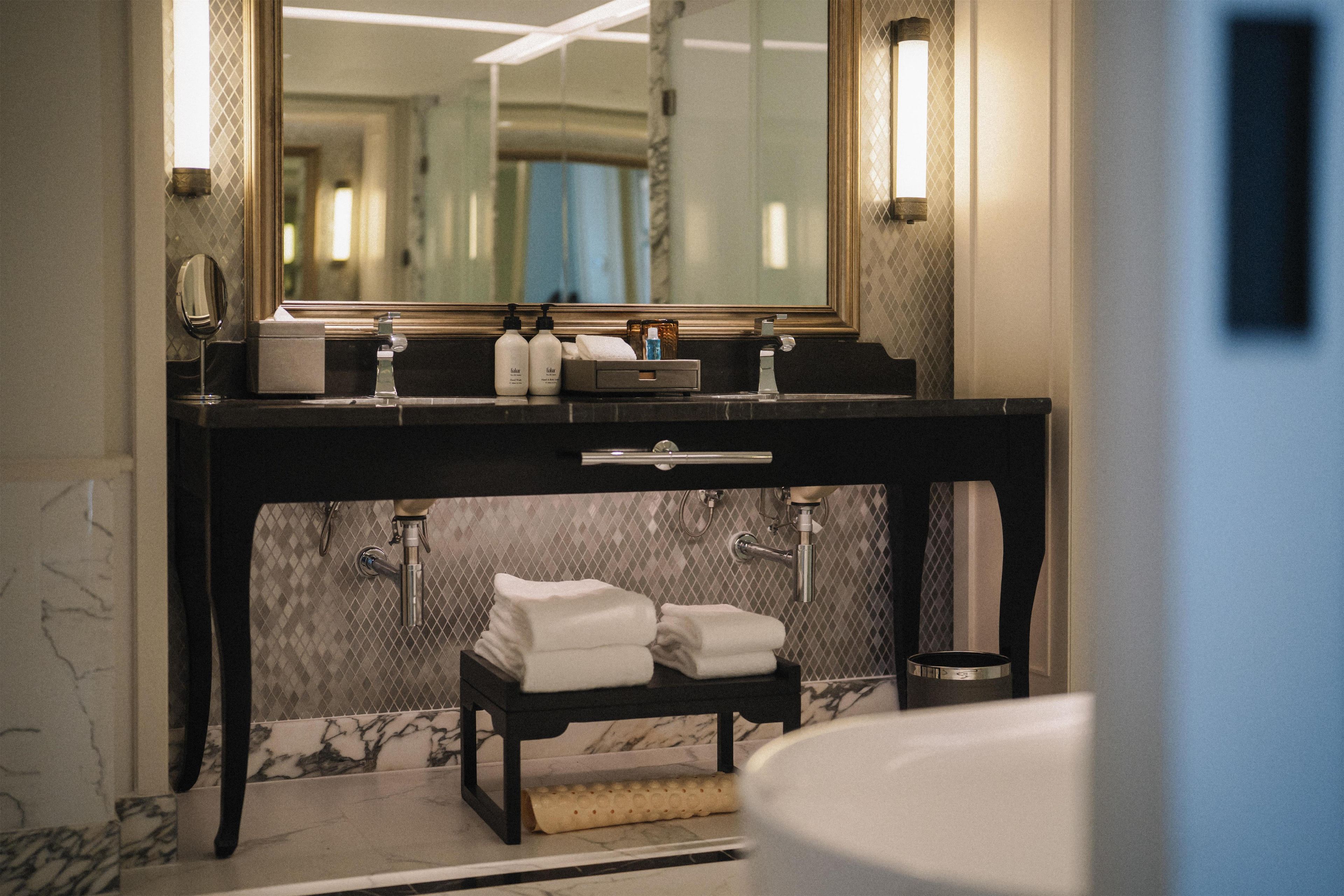 Un baño con suelo de mármol, tocador negro con espejo, toallero y lavabo.
