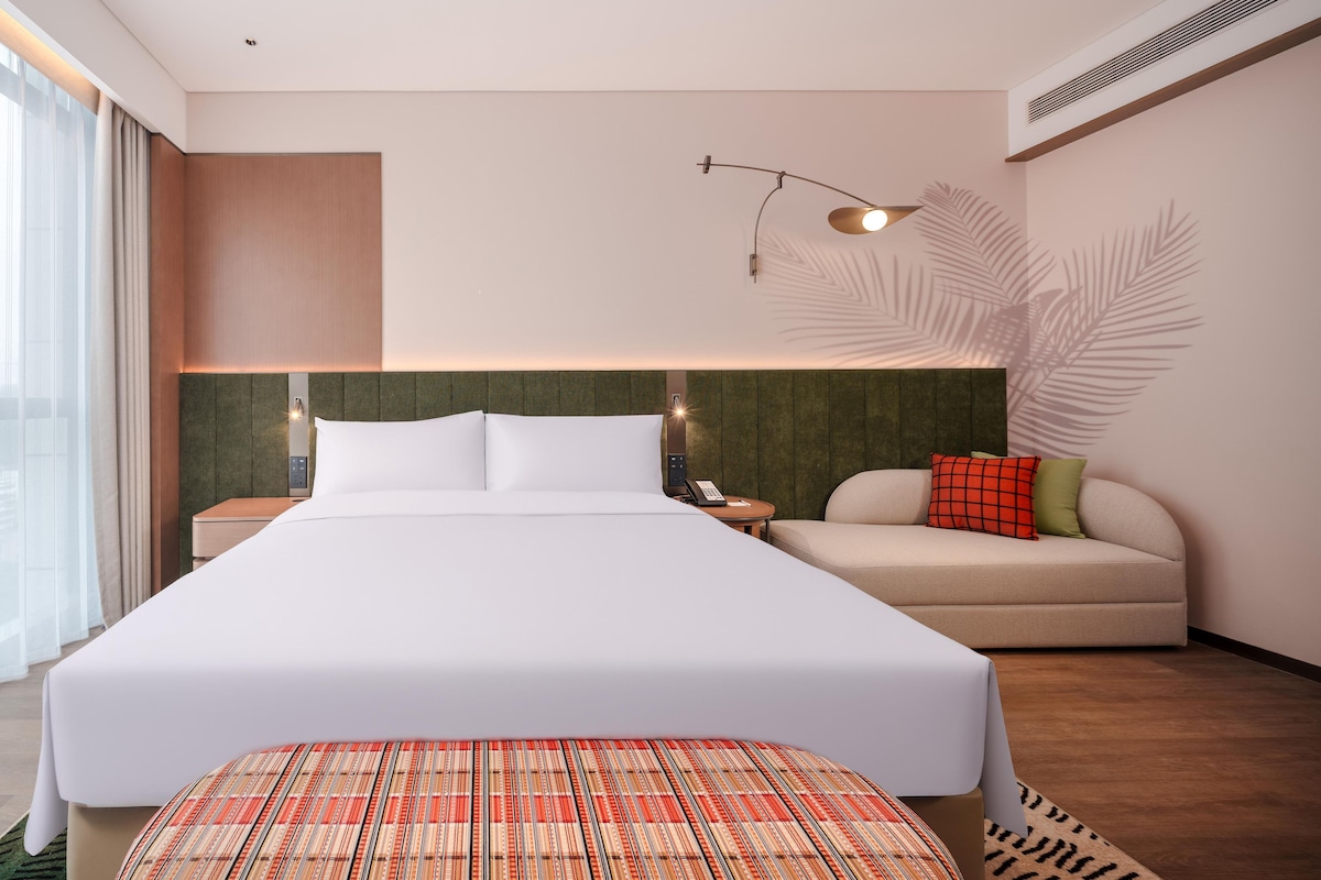 A modern hotel room with a large bed, a sofa, a nightstand with a phone, and a wall-mounted lamp.