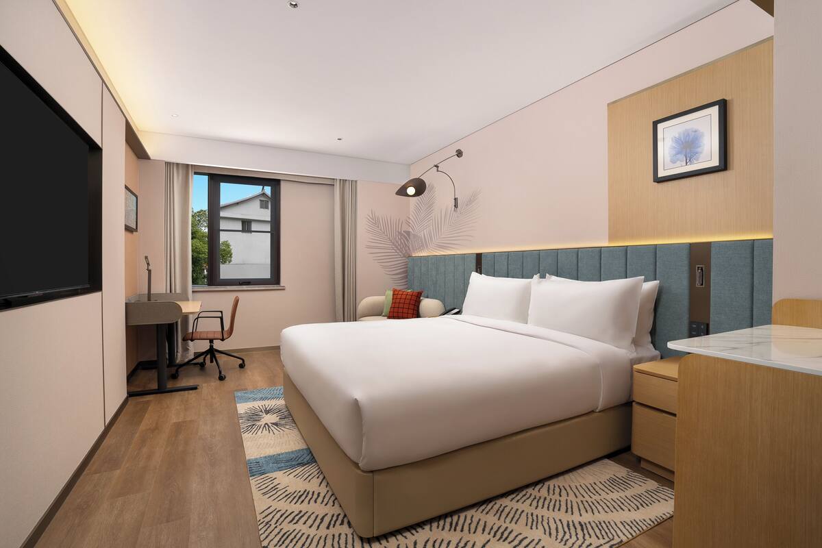 A modern hotel room with a large bed, a desk, a chair, a TV, and a window with a view of buildings.