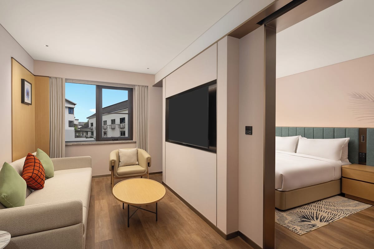 A modern hotel room with a sofa, armchair, round table, and a bed with a headboard.