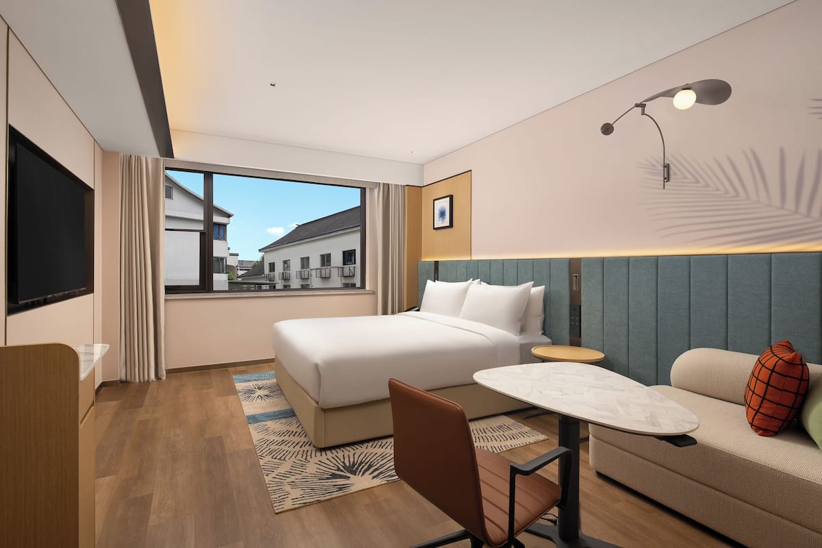 A modern hotel room with a large bed, a desk, a sofa, and a view of buildings outside the window.