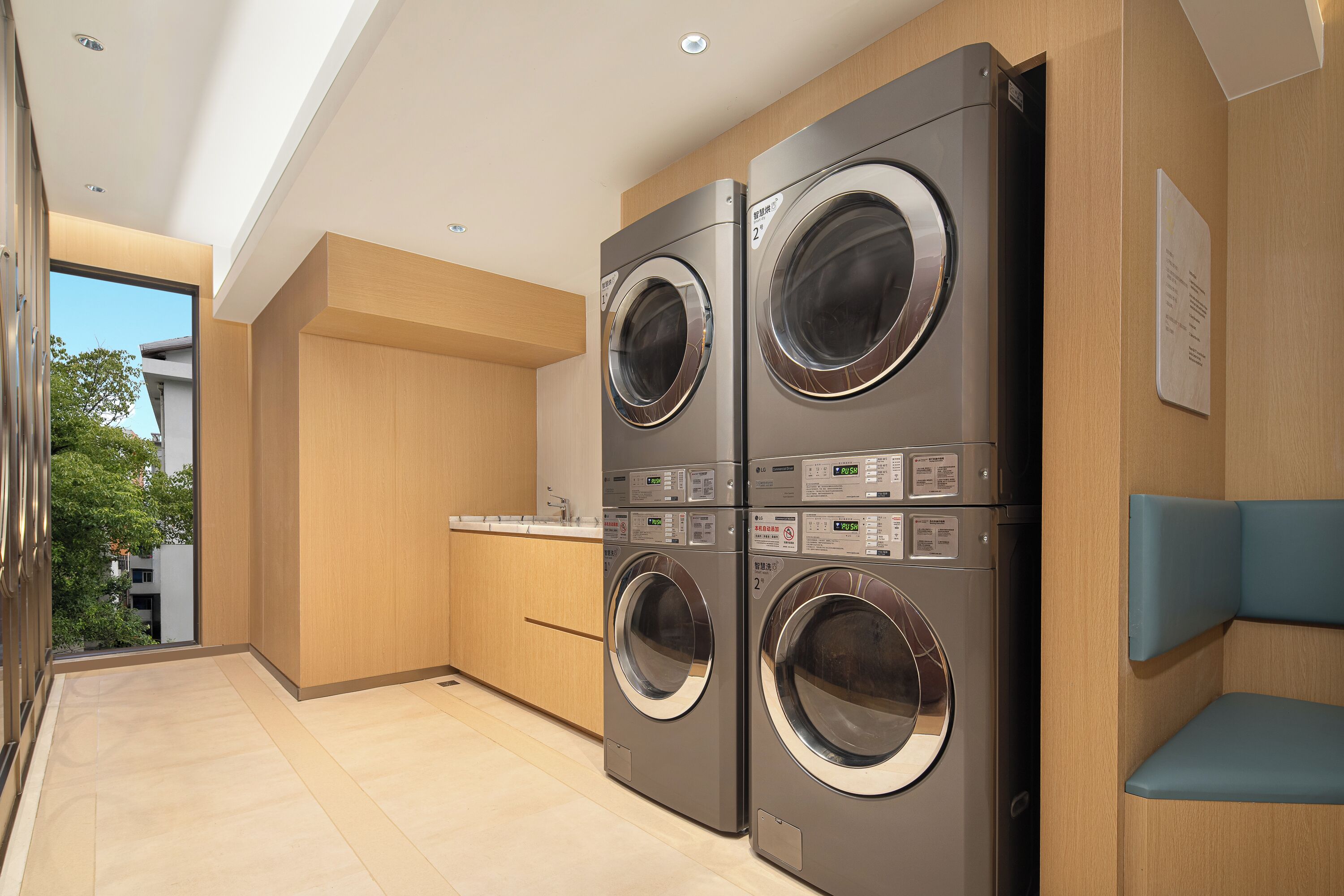A modern laundry room with two stacked front-loading washing machines, wooden cabinetry, and a view of the outdoors.