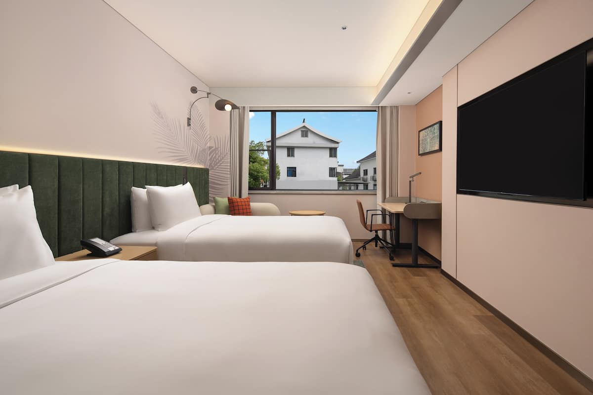 A hotel room with two beds, a desk, a chair, a TV, and a window with a view of buildings.