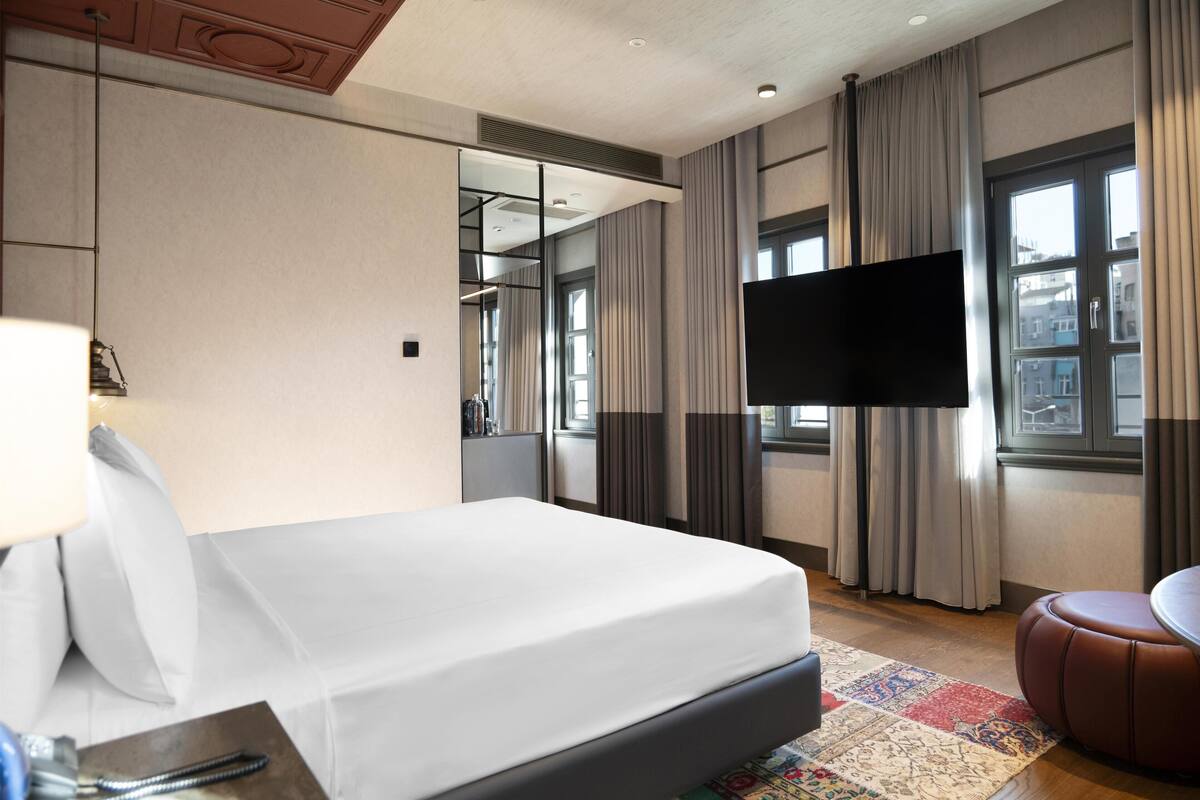 Une chambre d’hôtel dotée d’un grand lit, d’un téléviseur à écran plat fixé au mur, d’une fenêtre avec des rideaux et d’un petit coin salon avec un pouf en cuir.
