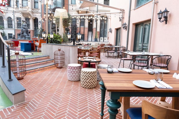 An outdoor patio area with brick flooring, wooden tables, and chairs, surrounded by buildings and a fountain.
