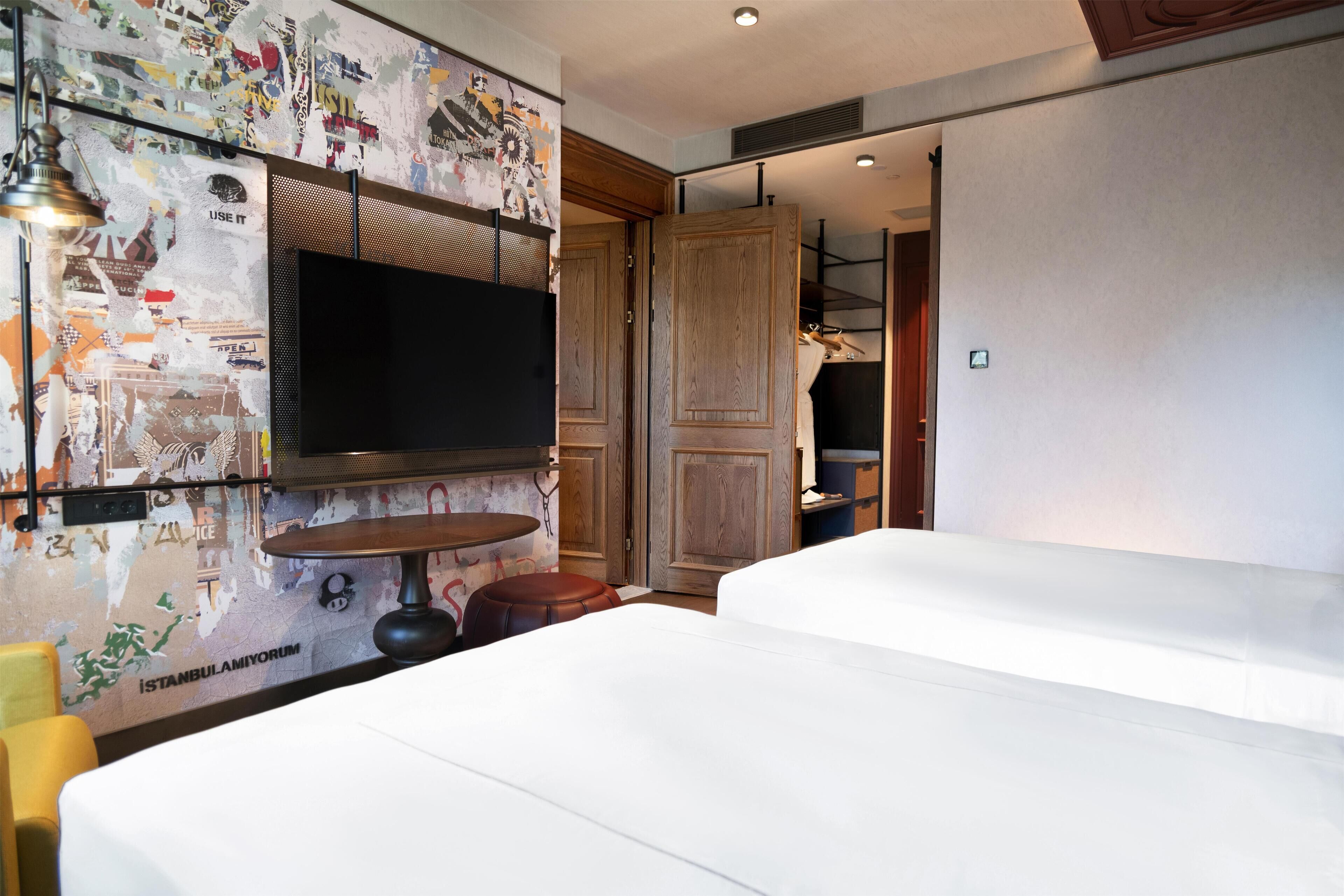 A hotel room with a large flat-screen TV mounted on a wall with a cityscape mural, a wooden door, and a white bed.