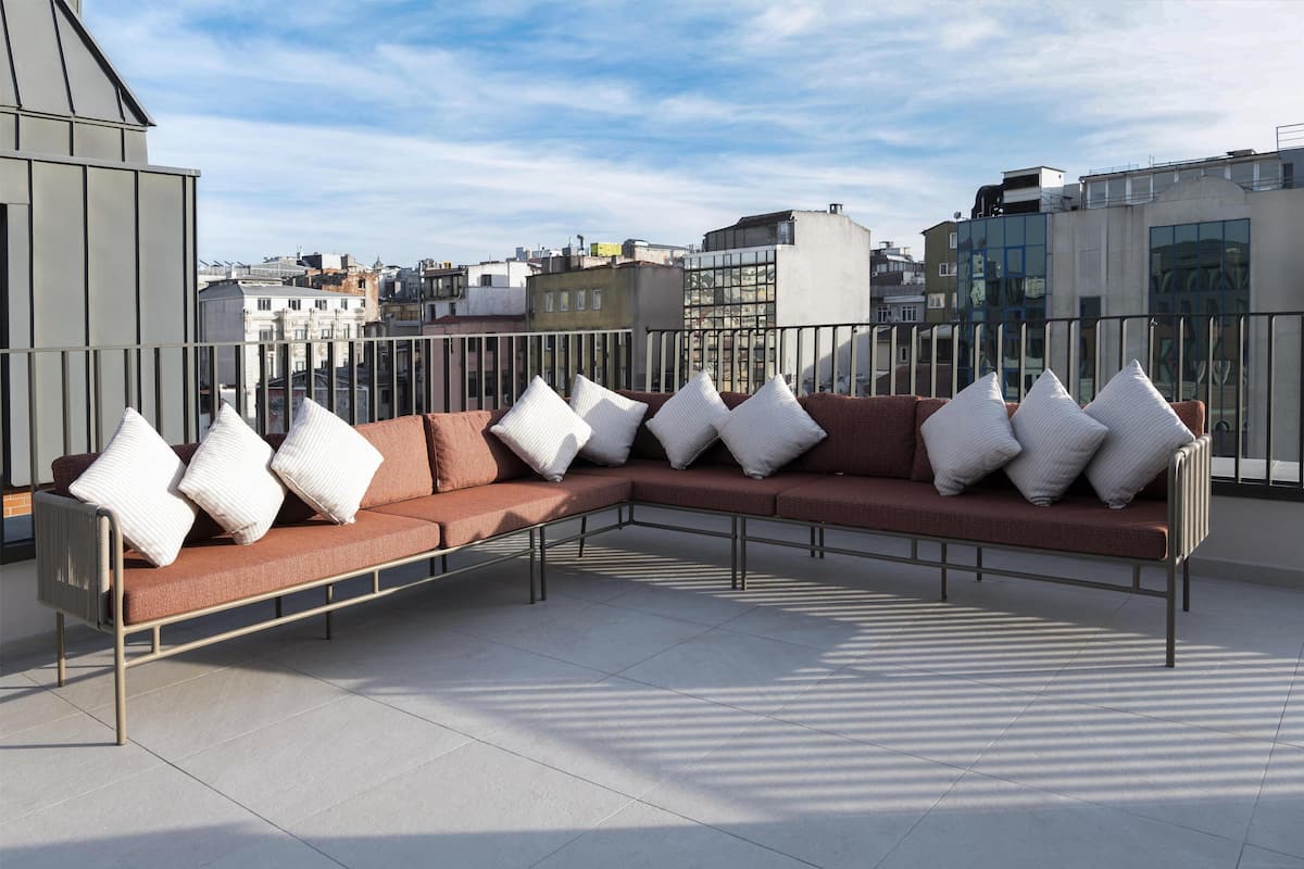 Un espace extérieur moderne aménagé avec un canapé et des coussins, une rambarde en métal et une vue sur le paysage urbain en arrière-plan.