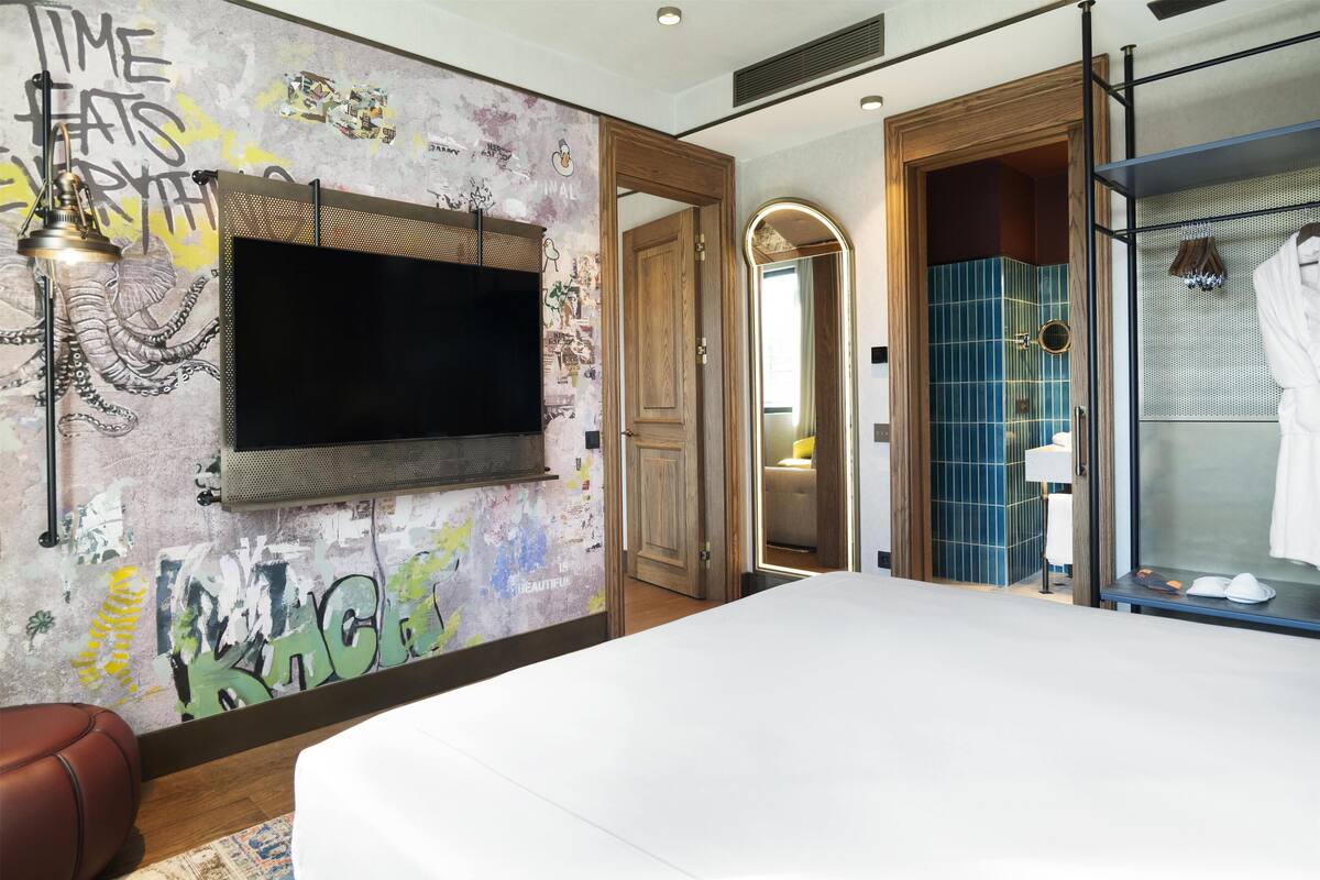 Une chambre d’hôtel moderne dotée d’un grand lit, d’un téléviseur à écran plat fixé au mur et d’une salle de bain avec une douche carrelée de carreaux bleus.