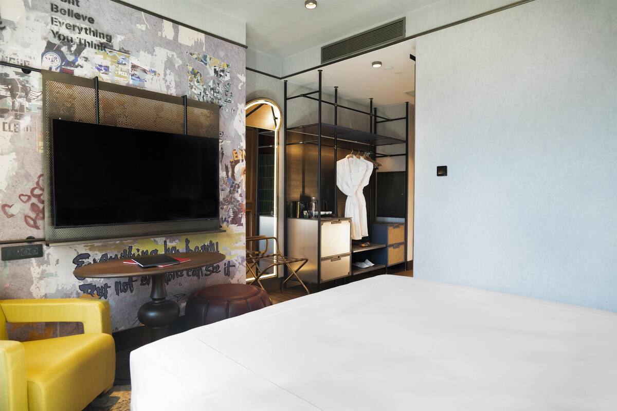 Une chambre d’hôtel moderne dotée d’un grand lit, d’un téléviseur à écran plat fixé au mur, d’un coin salon avec un fauteuil et un pouf, et d’un dressing où sont rangés un peignoir et une serviette.