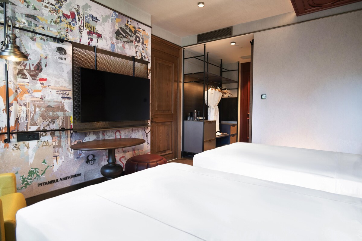 A hotel room with a large flat-screen TV mounted on a wall with a cityscape mural, two beds, a small round table, and a mirror.
