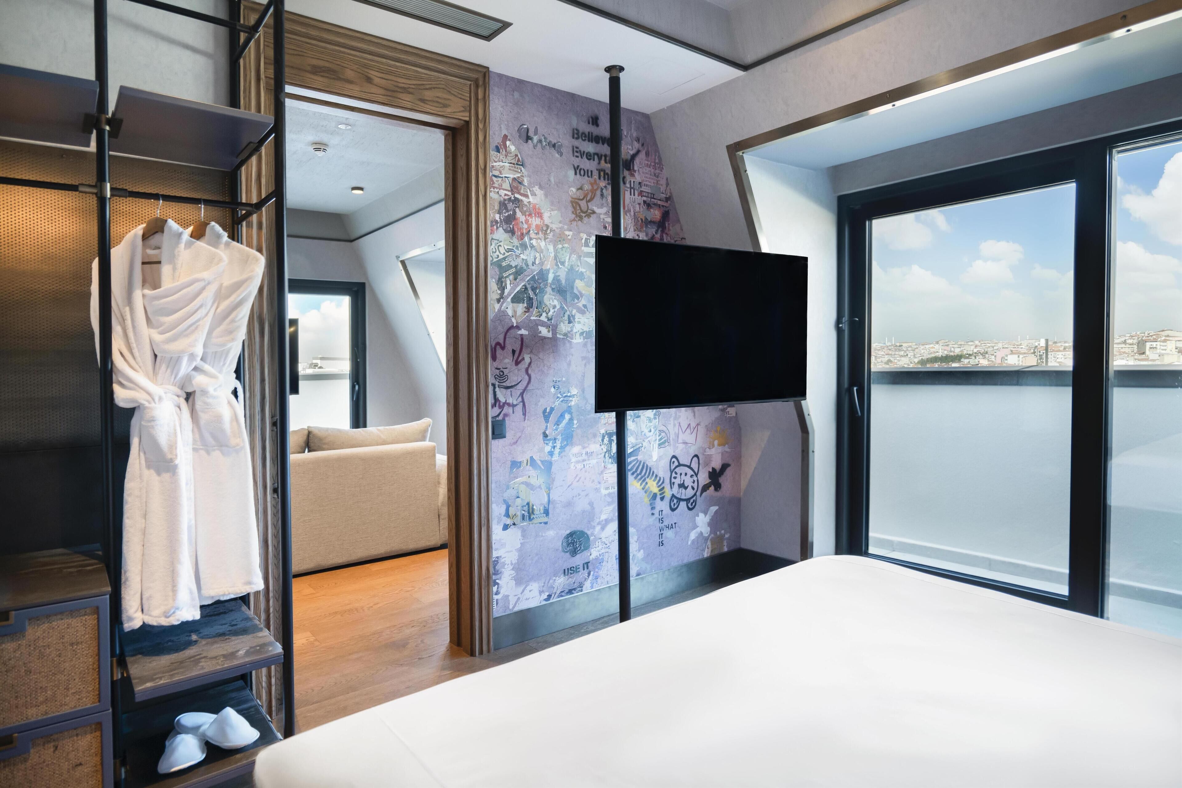 Una habitación de hotel moderna con una cama grande, un televisor montado en la pared, un balcón con vistas a la ciudad y un armario con batas.