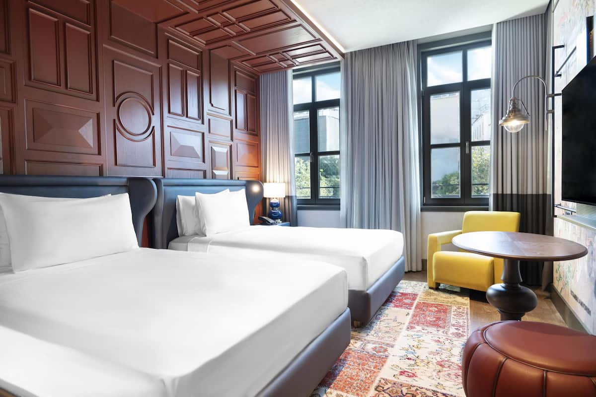 Une chambre d’hôtel avec deux lits, une petite table ronde, un fauteuil jaune et une grande fenêtre avec des rideaux.