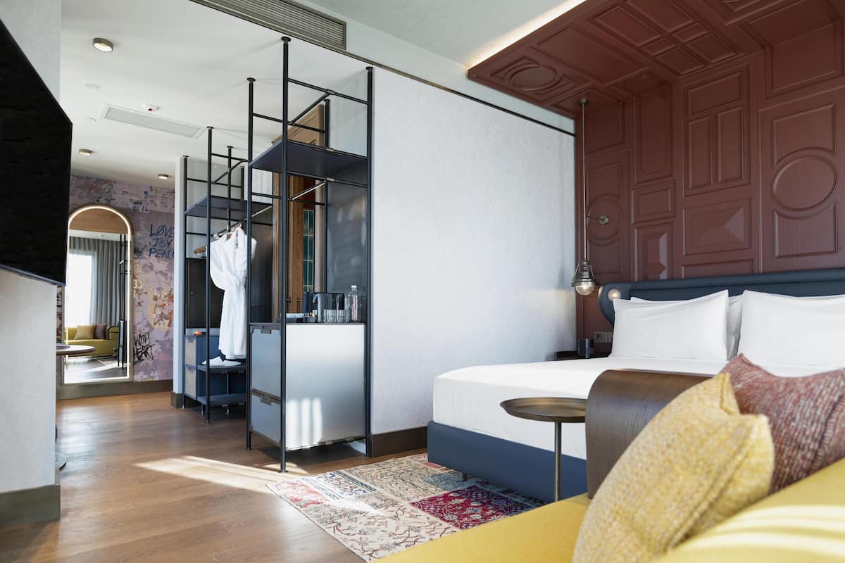 Une chambre d’hôtel moderne, dotée d’un grand lit, d’une petite table ronde, d’une armoire et d’une salle de bain visible à travers une paroi en verre.