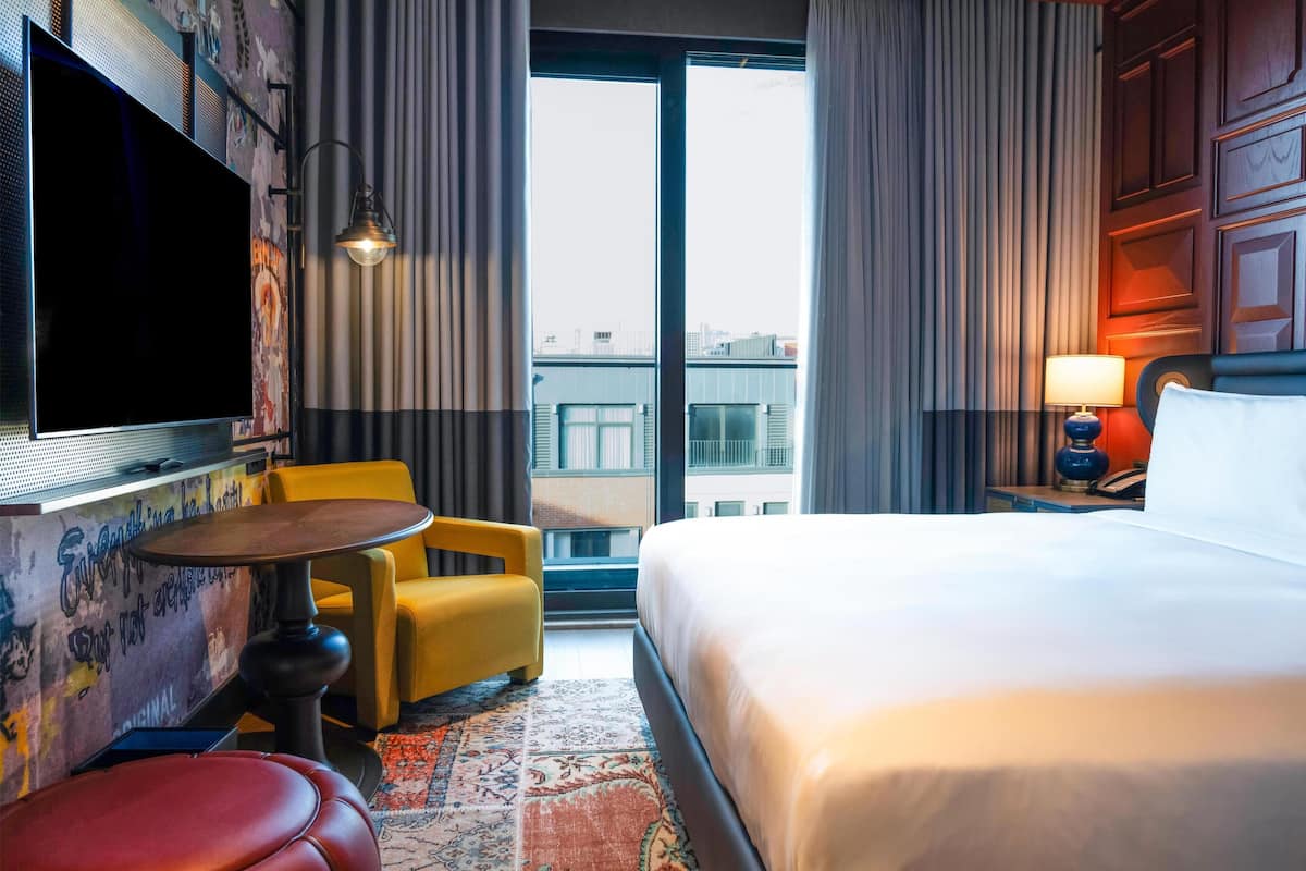 Une chambre d’hôtel avec un lit, une télévision, une petite table, une chaise jaune et une vue sur la ville grâce à de grandes fenêtres.