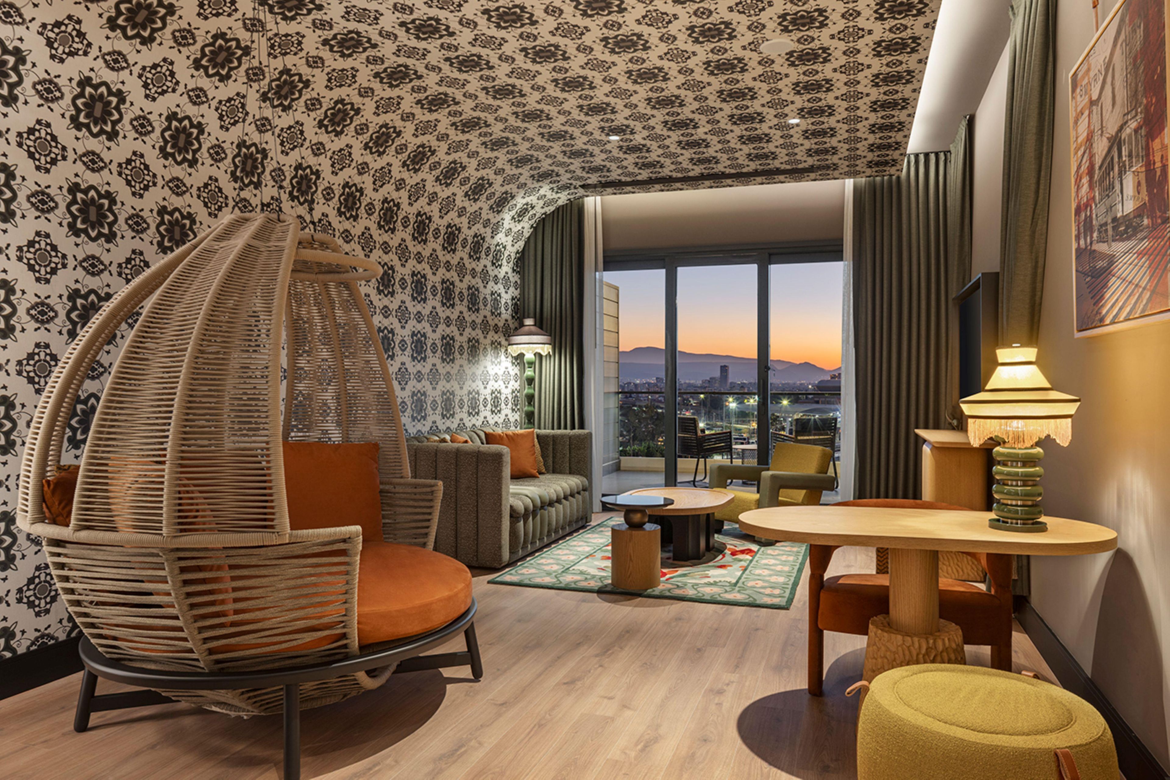 Ein modernes Hotelzimmer mit einem gebogenen Hängekorbsessel, einer Couch, einem runden Tisch und Blick auf die Skyline im Abendrot.