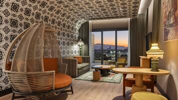 Ein modernes Hotelzimmer mit einem gebogenen Hängekorbsessel, einer Couch, einem runden Tisch und Blick auf die Skyline im Abendrot.