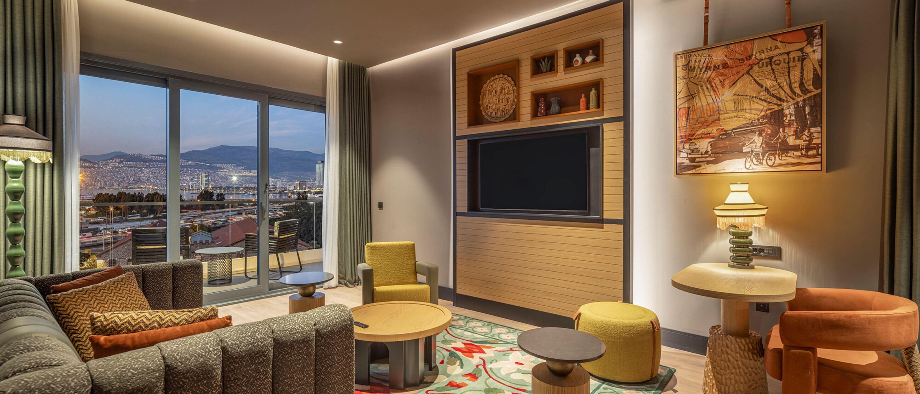 A modern living room with a large window offering a city view, a flat-screen TV mounted on a wooden panel, and a cozy seating area with a round coffee table.