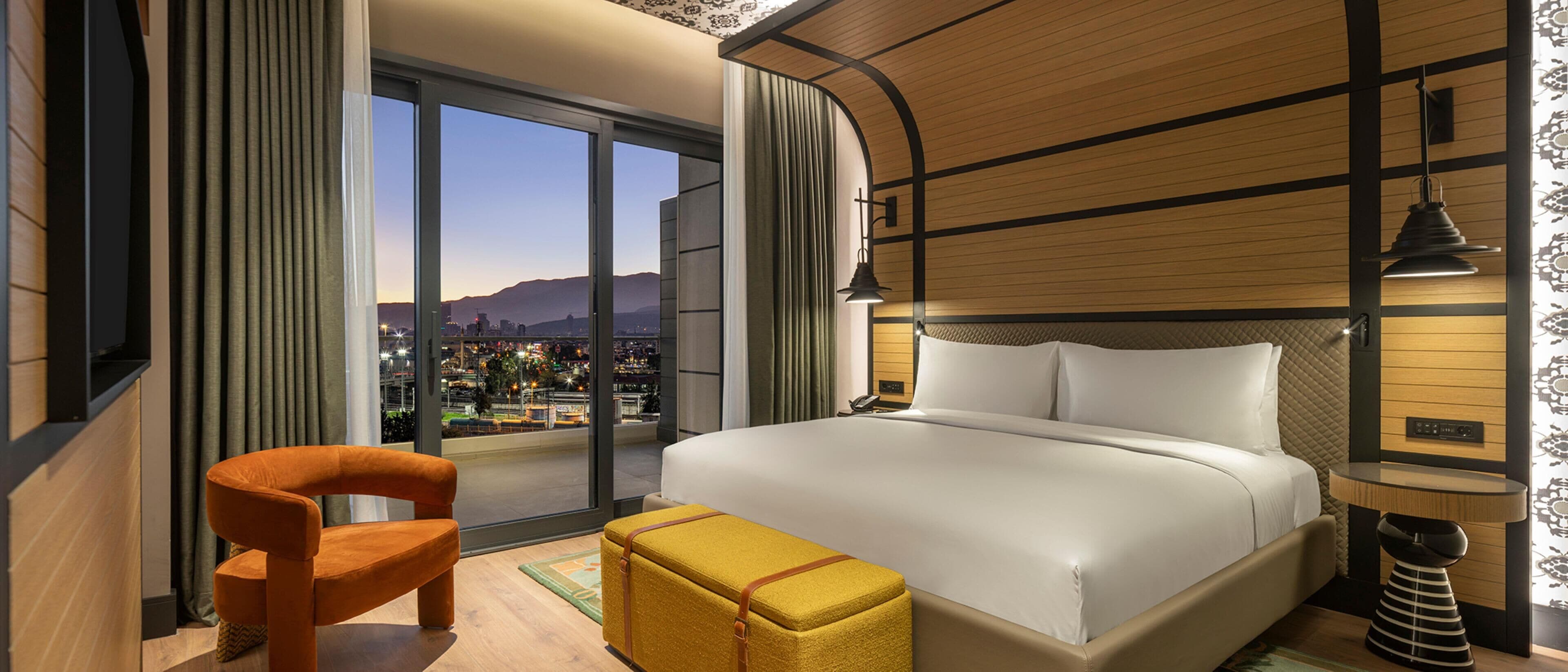 Habitación de hotel con una cama grande, una silla, una mesita y vistas a un paisaje urbano a través de un ventanal amplio.
