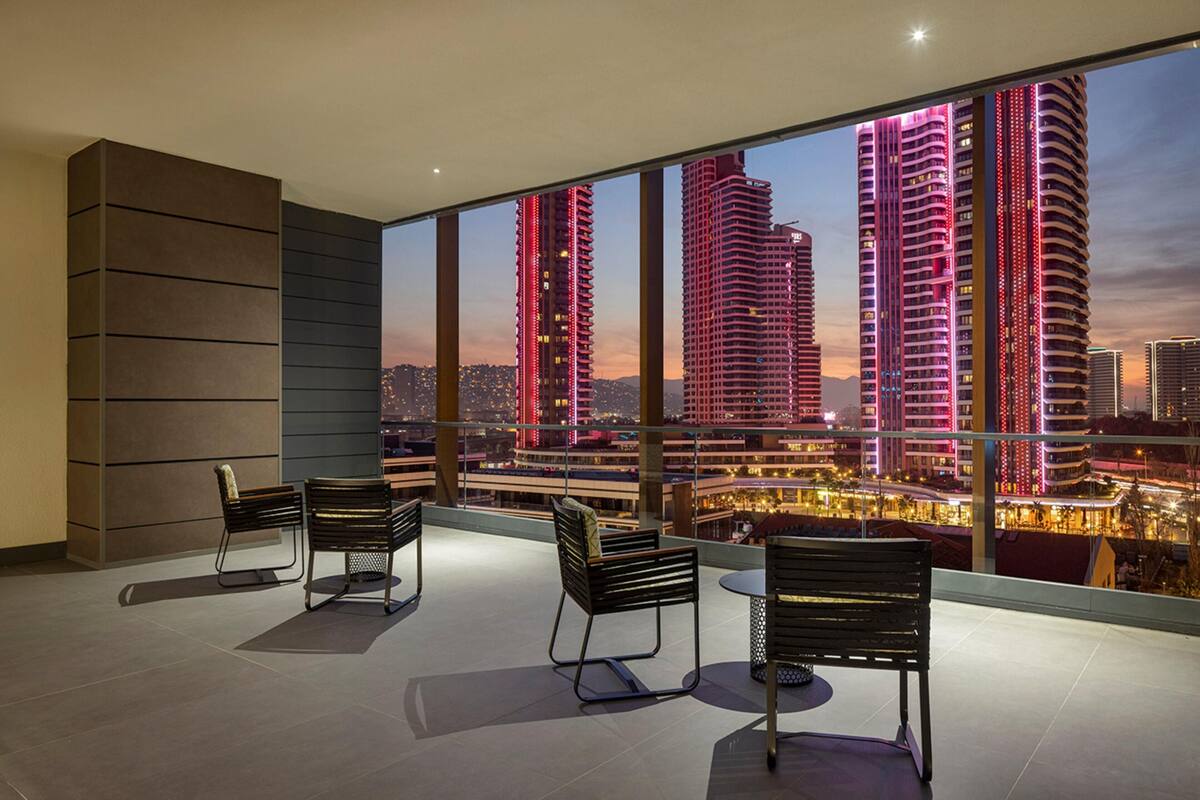 Une chambre d’hôtel moderne dotée d’un grand balcon, de deux chaises noires et offrant une vue sur les gratte-ciel illuminés au crépuscule.