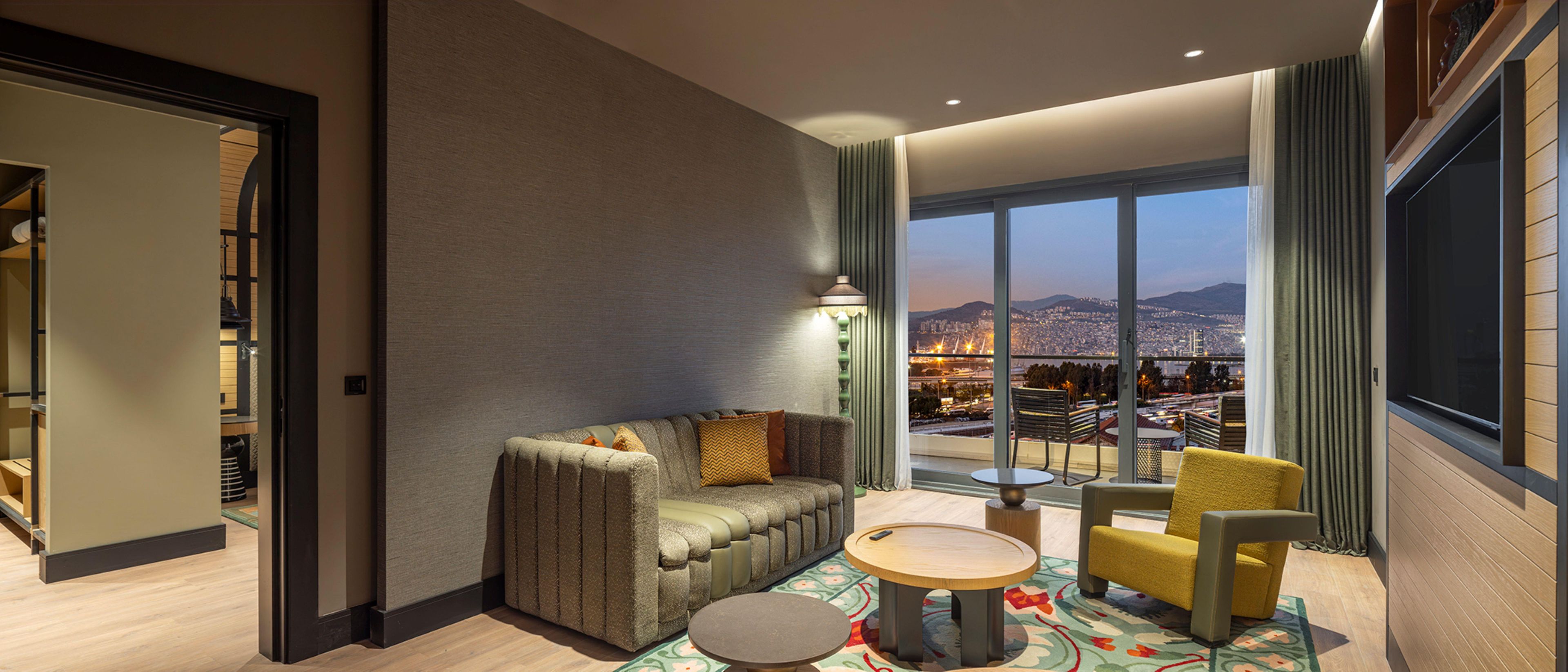 A modern hotel room with a sofa, armchair, and round table, featuring a large window with a city view.