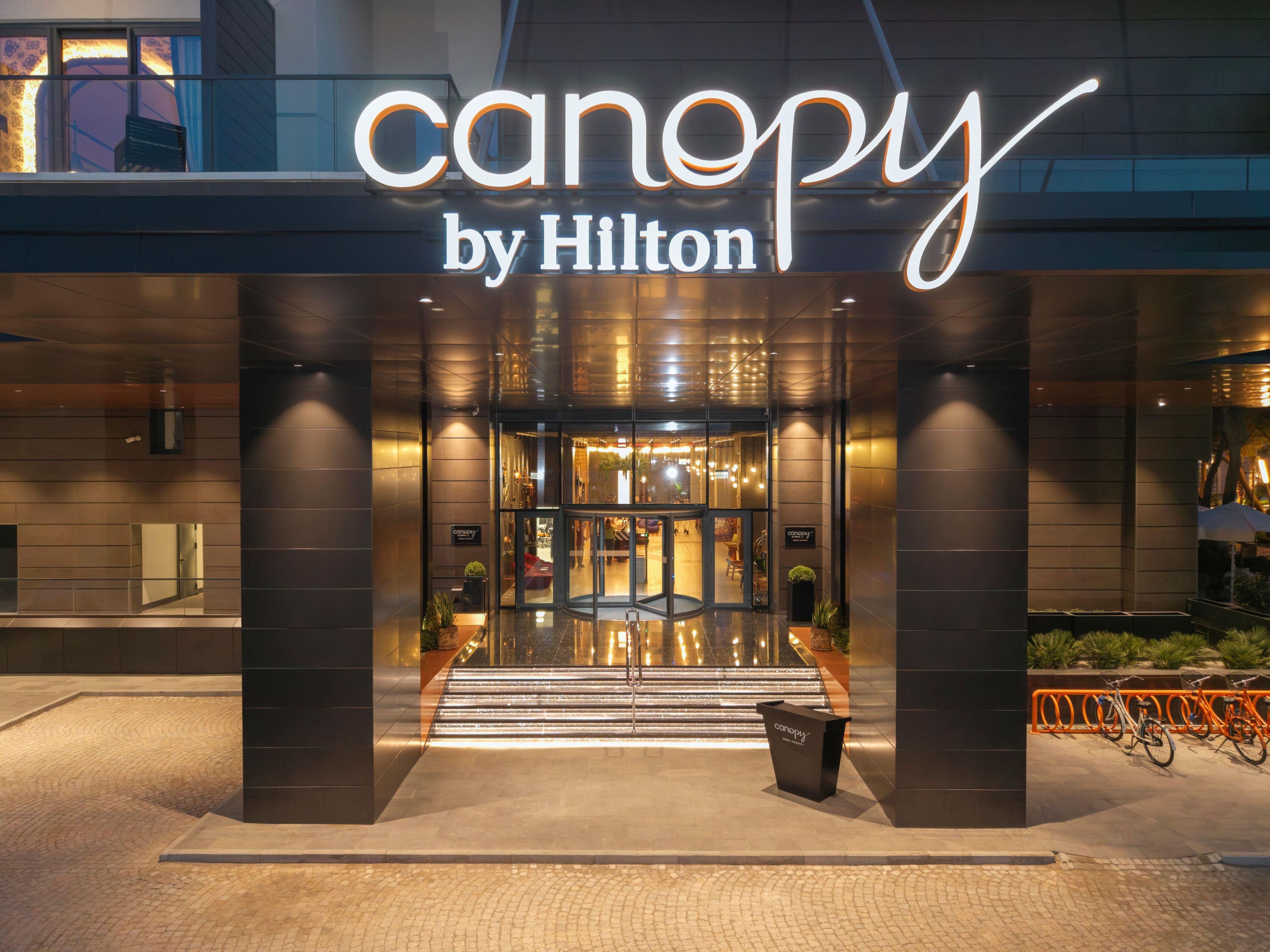 Der Eingang eines Hotels mit modernem Design, der eine Glasfassade und ein Schild mit der Aufschrift 'canopy by Hilton' zeigt.