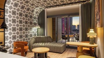 A modern hotel room with a patterned wallpaper, a sofa, two armchairs, a small round table, a lamp, and a view of a cityscape through a large window.