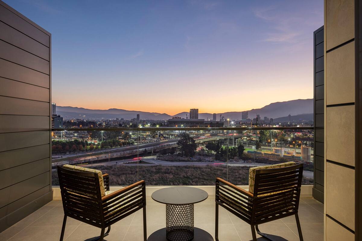 Un balcon avec deux chaises et une table, d’où l’on peut admirer un paysage urbain avec des montagnes en arrière-plan.