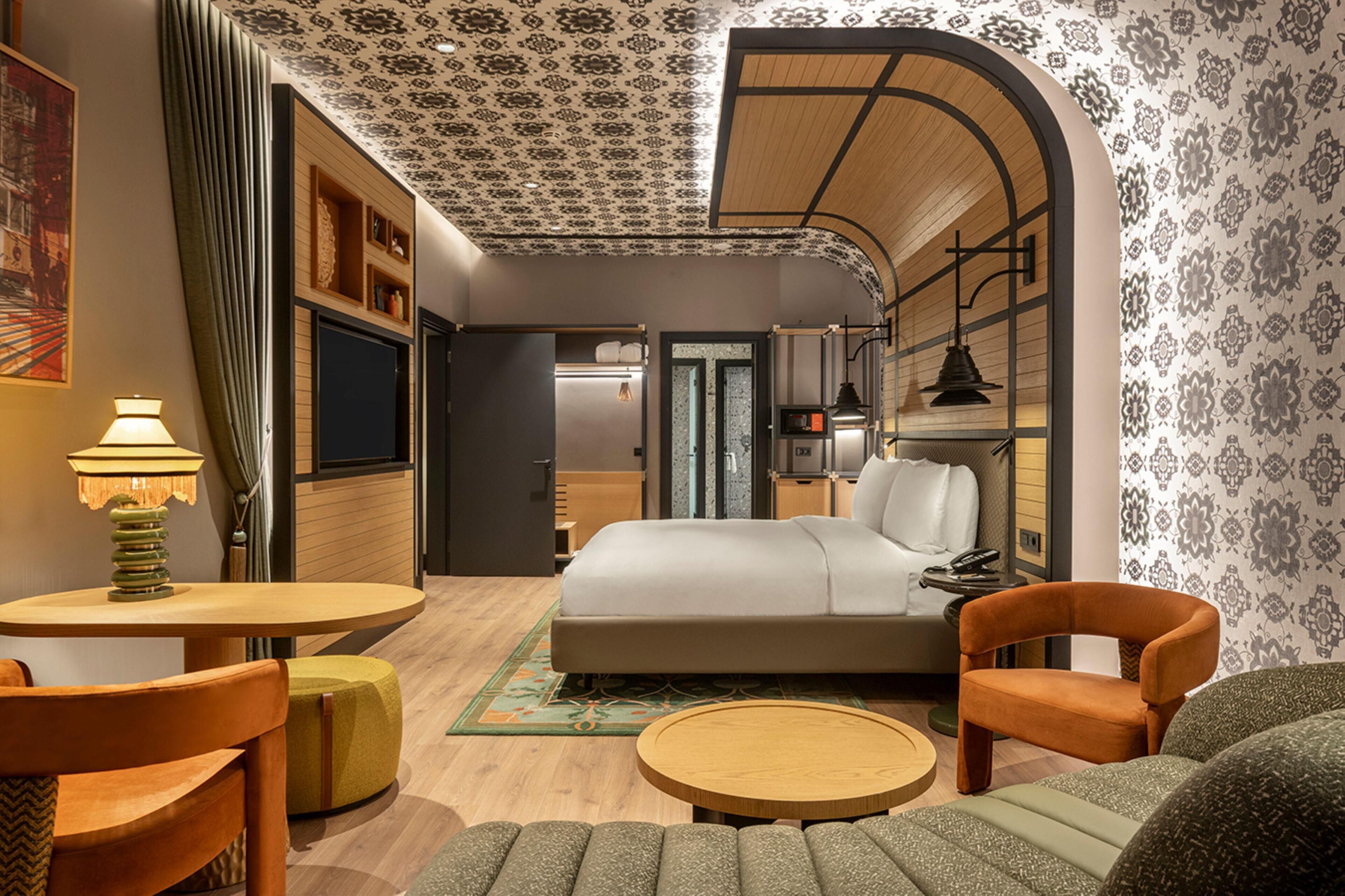 A hotel room with a bed, a round wooden table, two chairs, a small ottoman, a lamp, and a wall-mounted TV.