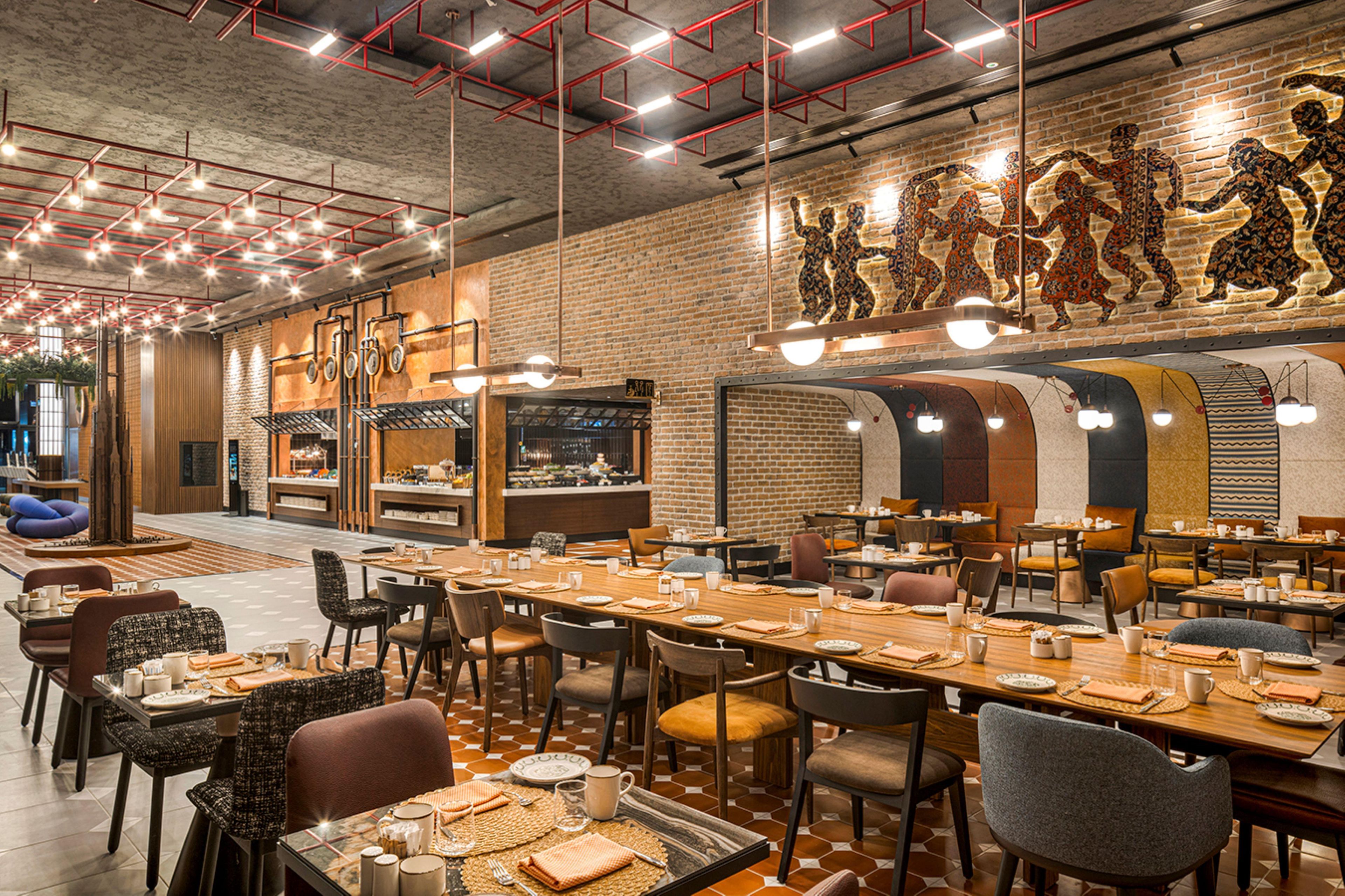 Un restaurante con paredes de ladrillo, luces colgantes y un mural de figuras danzantes. Las mesas están dispuestas con platos y cubiertos.