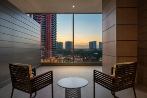 Un balcon avec deux chaises et une table, d’où l’on peut admirer un paysage urbain avec ses bâtiments et un coucher de soleil.