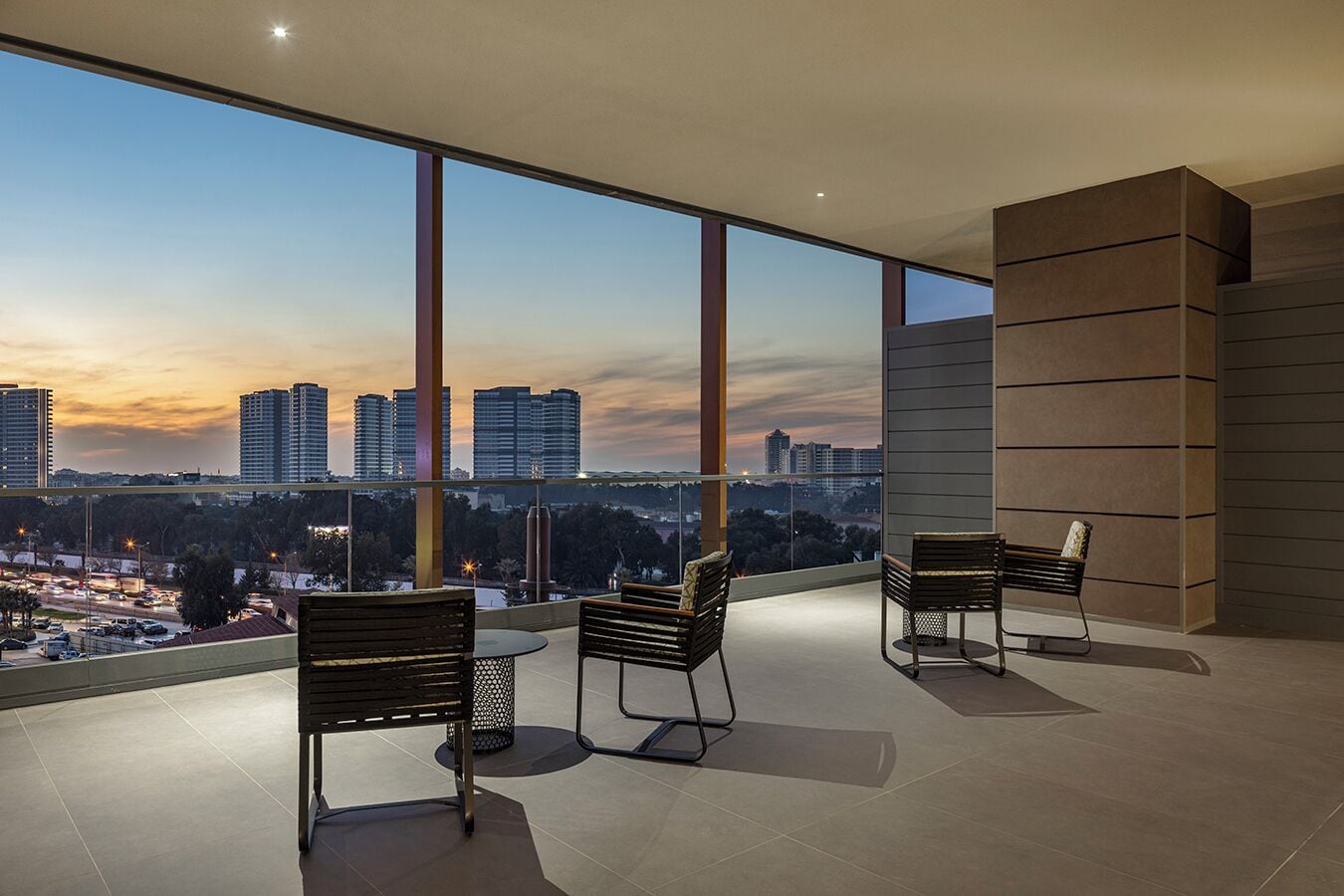 A modern balcony with large windows, two wooden chairs, and a view of a cityscape at dusk.