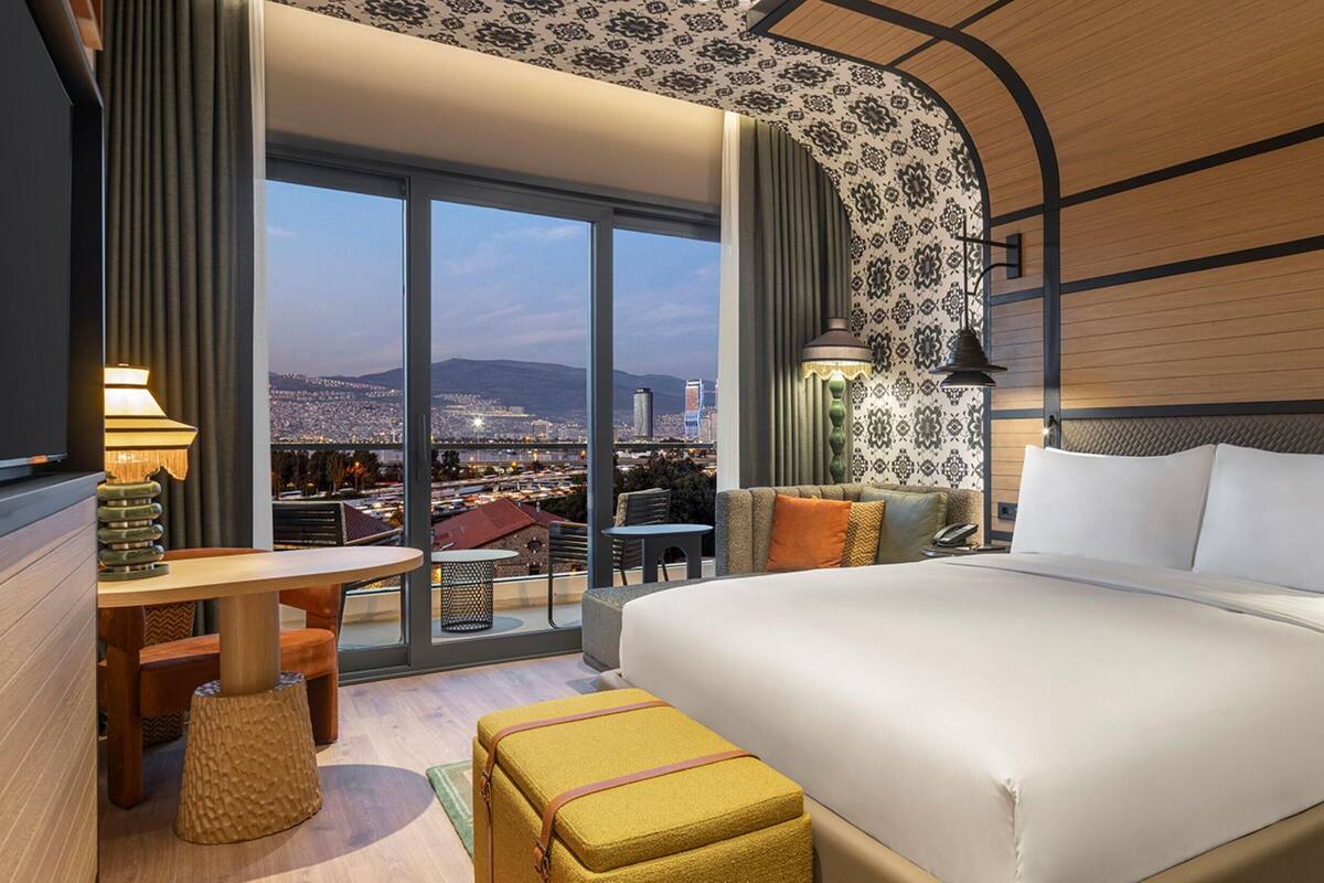 Une chambre d’hôtel avec un grand lit, une petite table, un canapé et une vue sur le paysage urbain grâce à de grandes fenêtres.