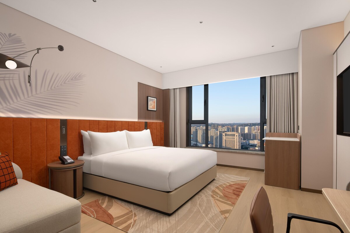 Habitación de hotel moderna con una cama grande, un escritorio, una silla, una mesita de noche, un teléfono, una ventana con vista a la ciudad y una lámpara de pared.