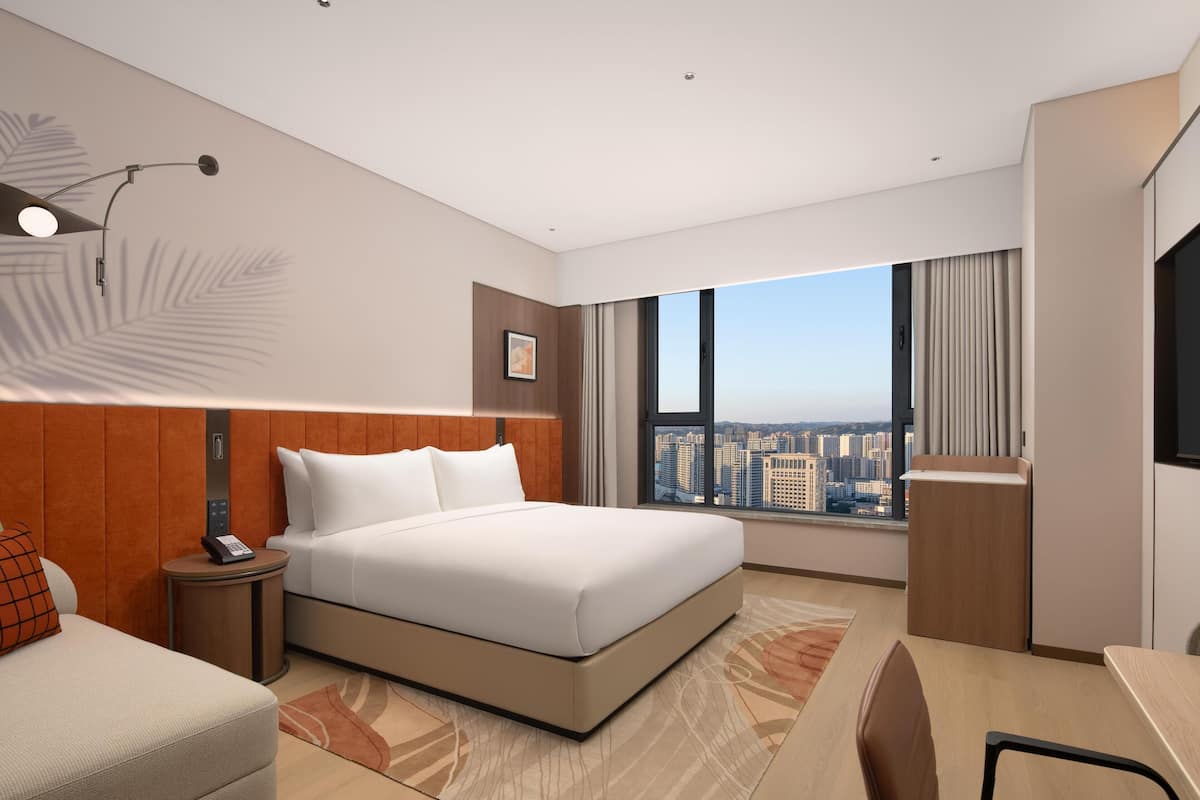A modern hotel room with a large bed, a desk, a chair, a nightstand, a phone, a window with a city view, and a wall-mounted light fixture.