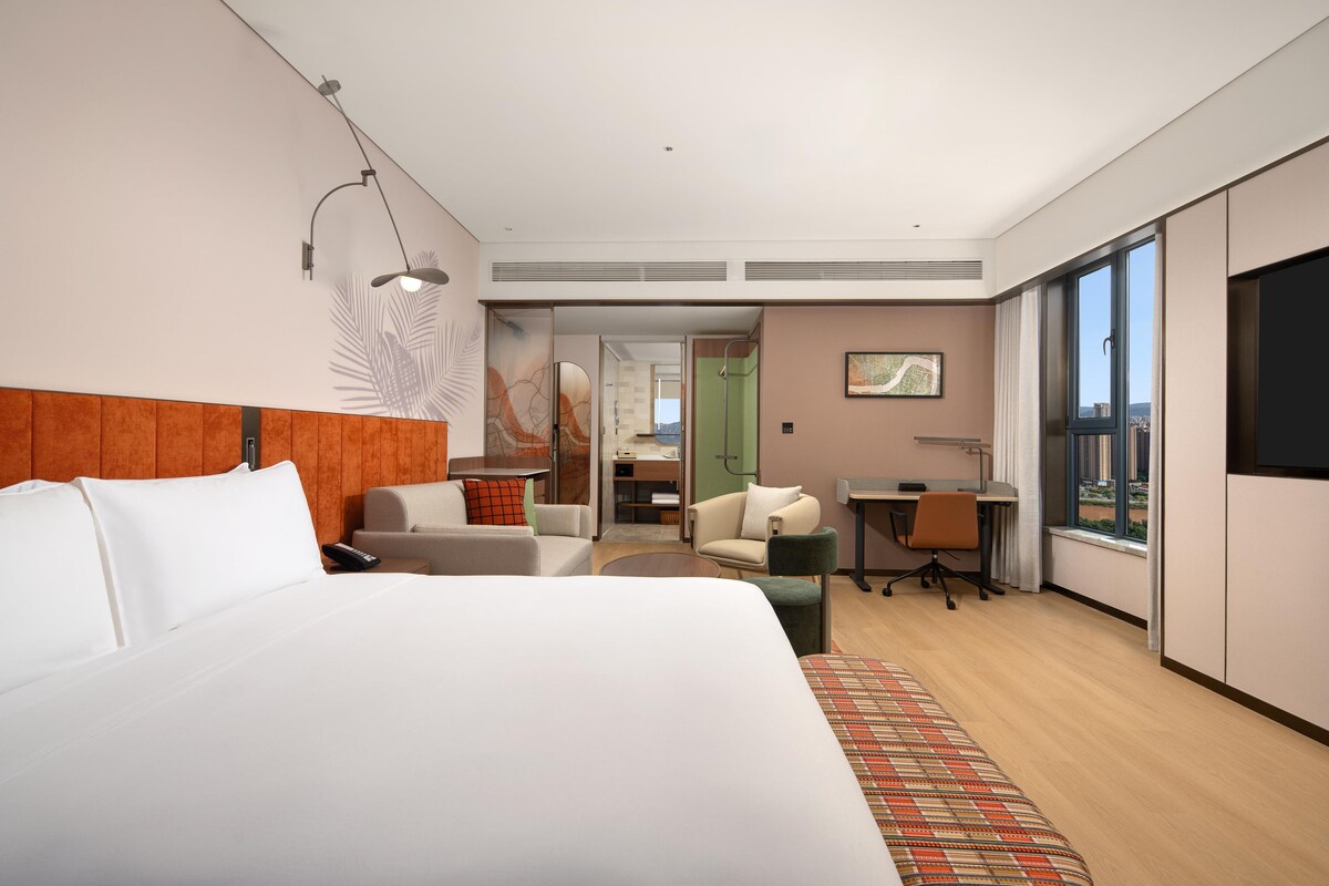 Habitación de hotel moderna con una cama grande, un escritorio, una silla y vistas a la ciudad desde la ventana.