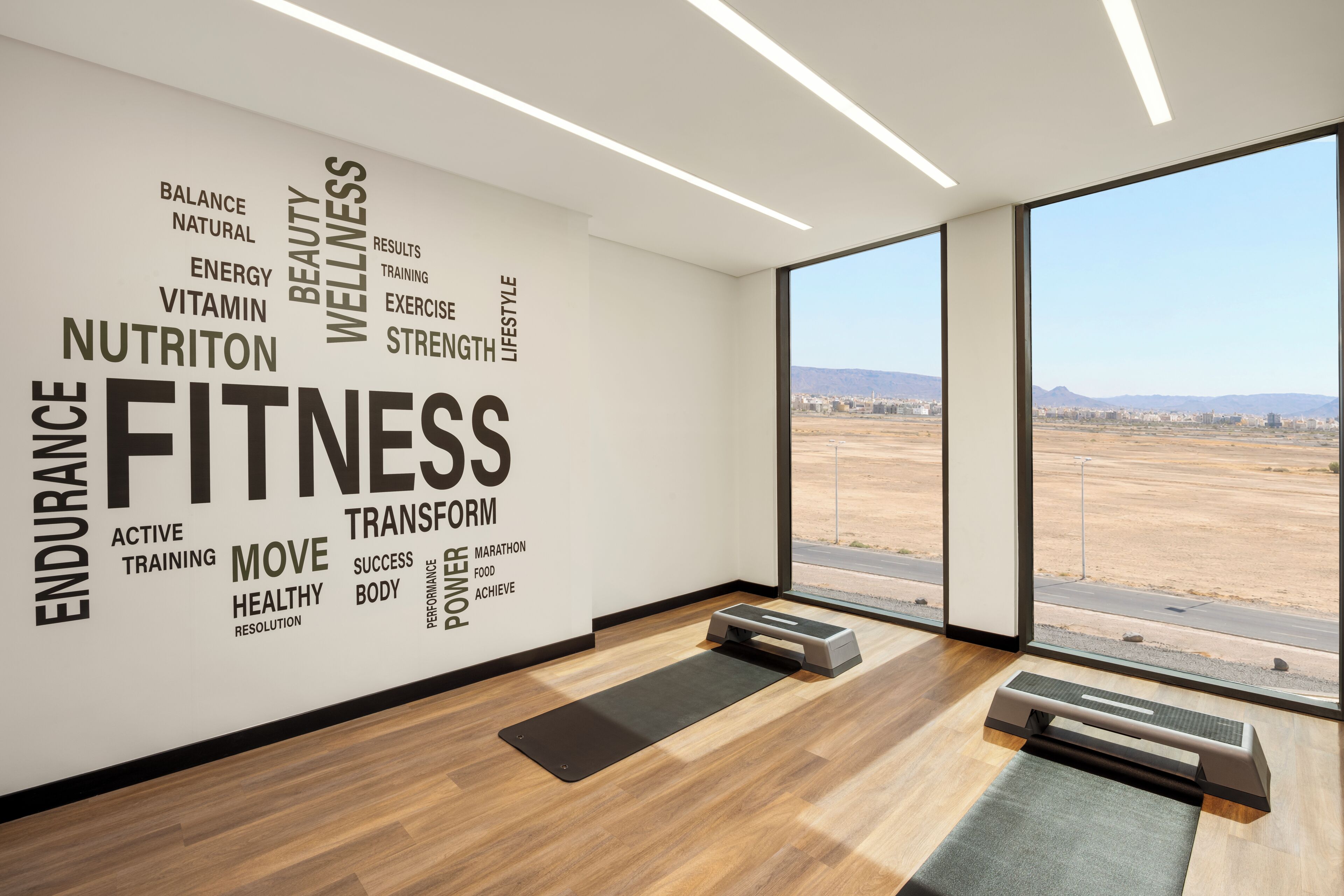 Un gimnasio moderno con una gran pared de palabras motivacionales, suelo de madera, un escritorio de pie y vistas a un paisaje desértico.